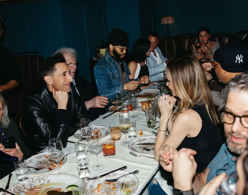 Adrien Brody seated at a long table with other guests at a restaurant, looking towards the right.