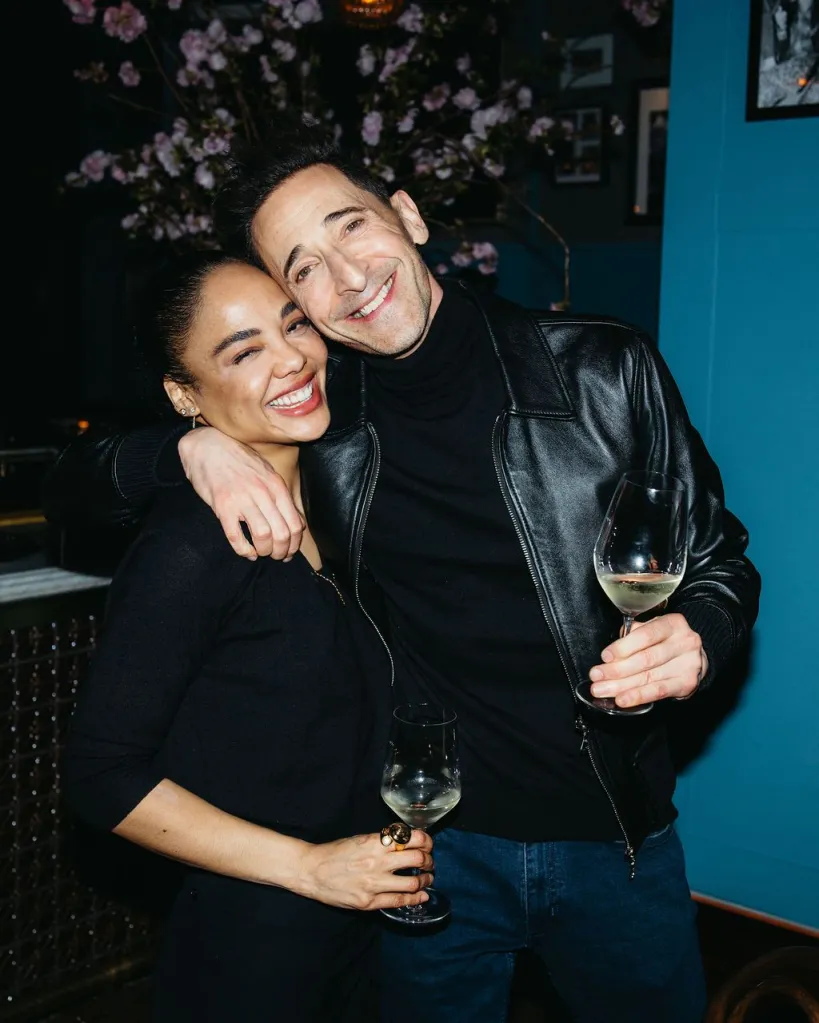 Adrien Brody and Georgina Chapman smiling and embracing, holding wine glasses.