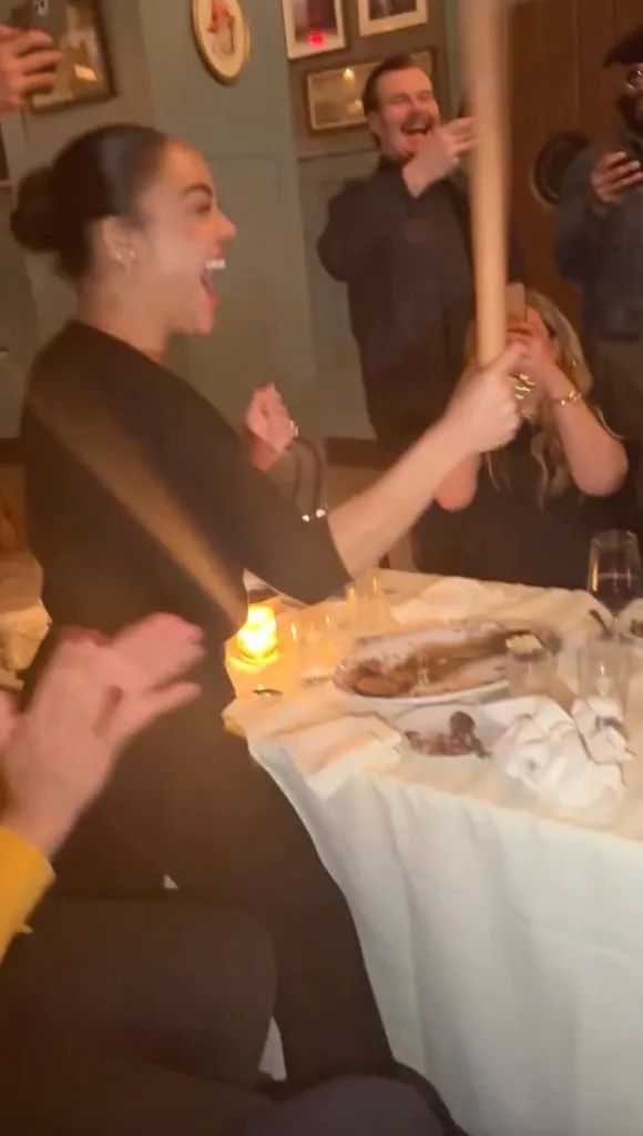A woman at a restaurant table, mid-laugh with her mouth open, holds a rolling pin over a plate of food.