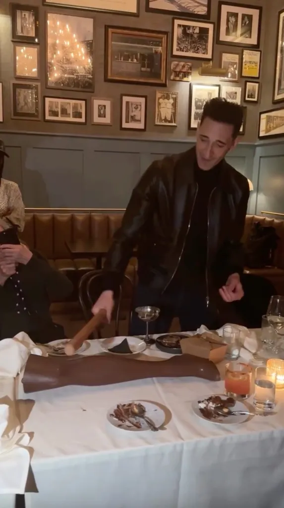 A man in a black leather jacket smashing a chocolate dessert shaped like a leg with a wooden hammer.