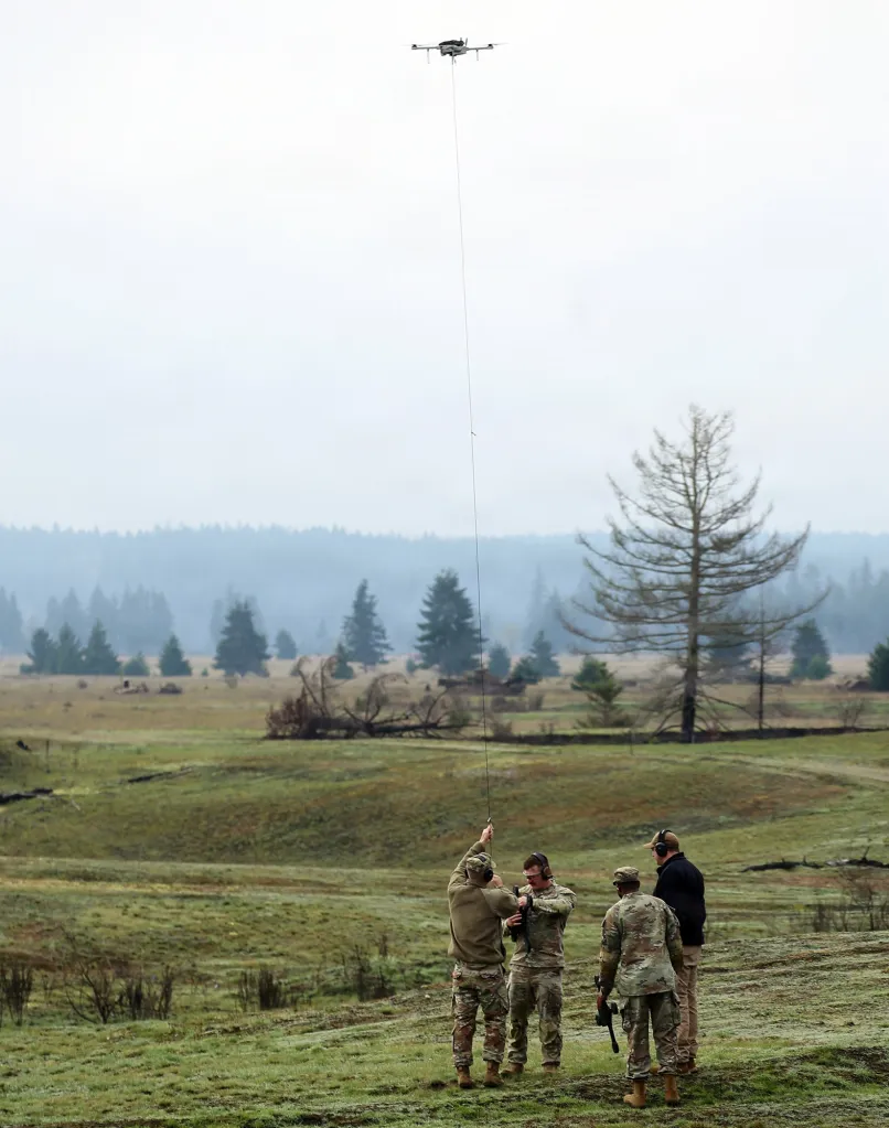 Military personnel operating a tethered drone in a field.