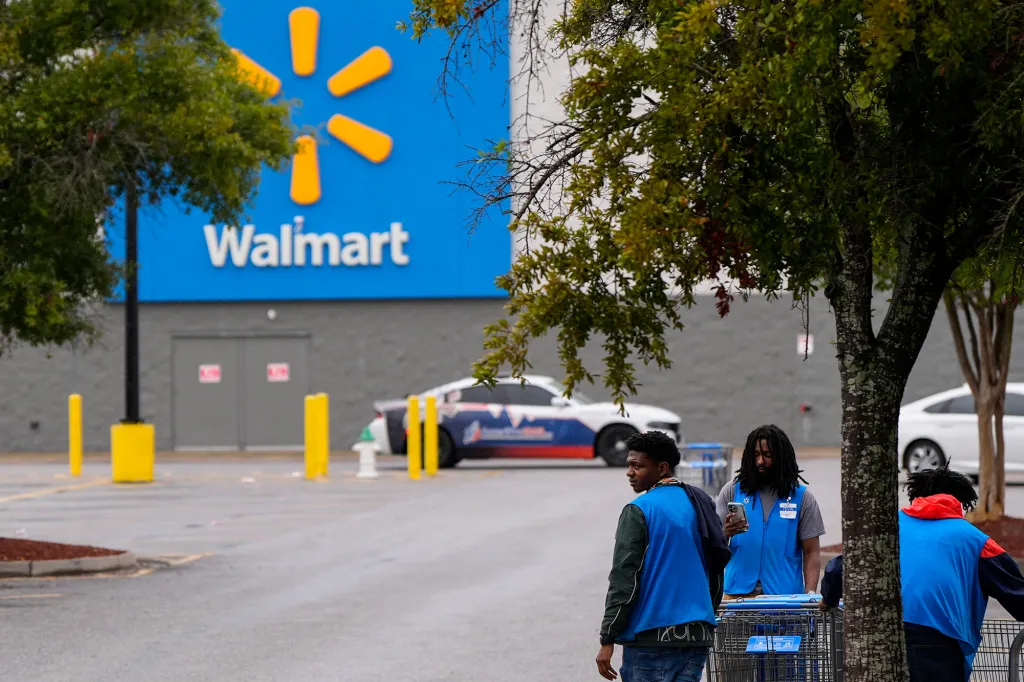 Walmart employees gather near shopping carts in the parking lot with the store entrance and logo visible in the background.