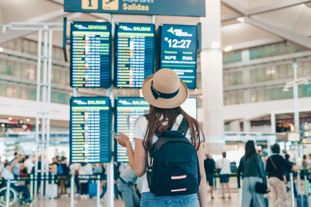 A young woman with a straw hat and backpack checking departure information at an airport.