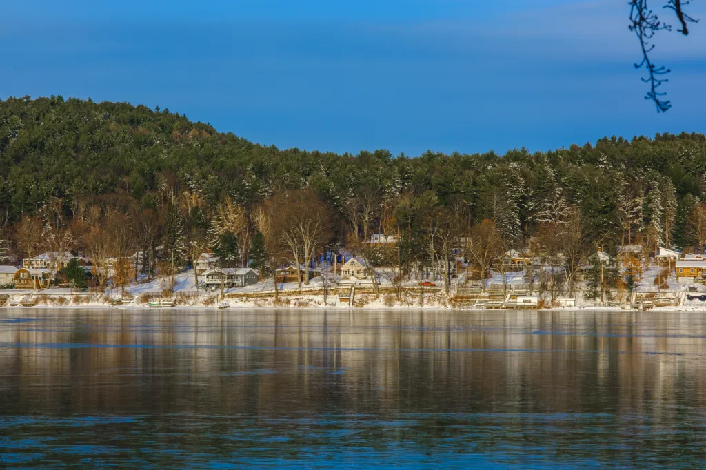 A winter landscape of Copake Lake, with snow-covered trees and houses on the shore reflecting in the still water under a clear blue sky.