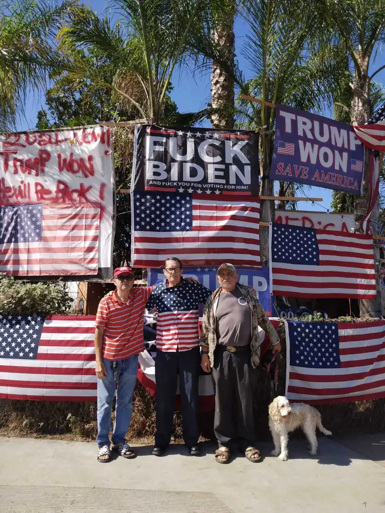 Kerry Sheron, center, with two other men and a dog standing in front of a house covered in pro-Trump signs and American flags.