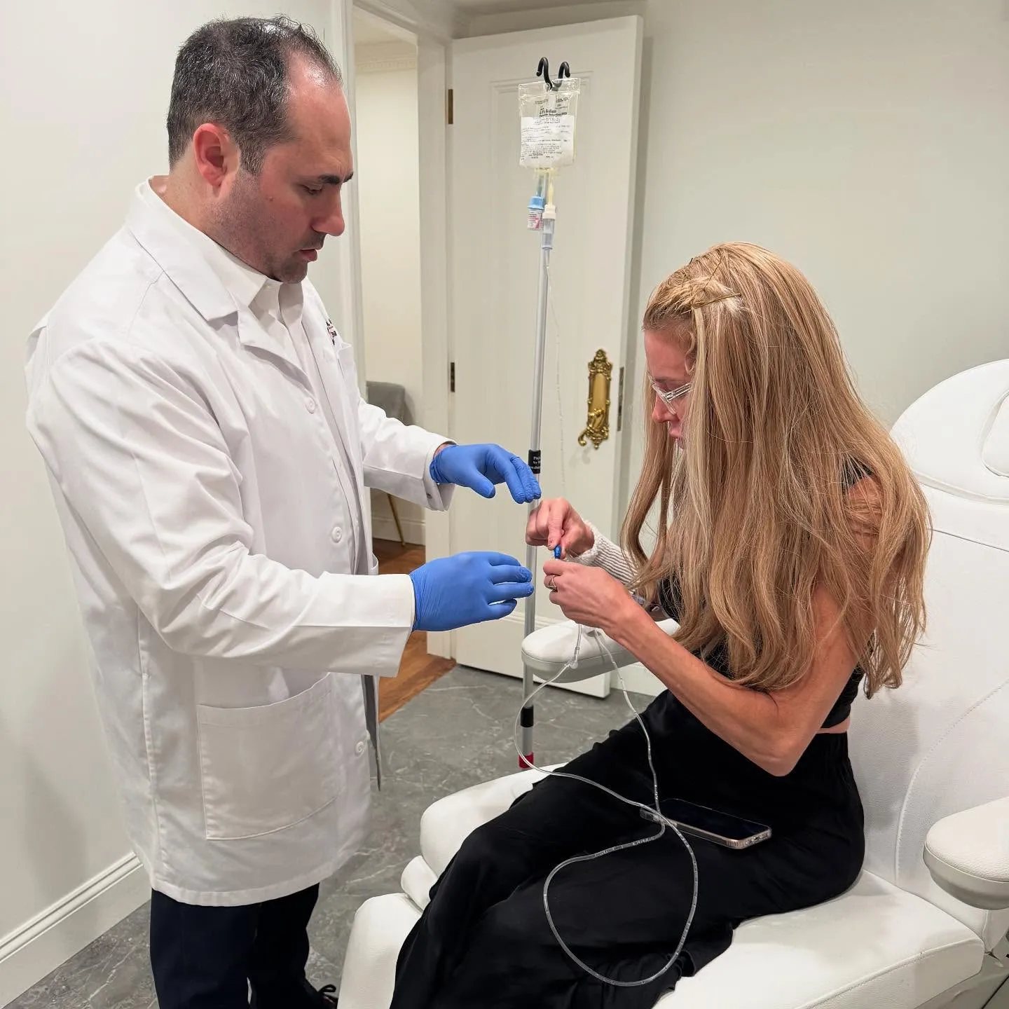 A doctor in a white coat and blue gloves prepares an IV drip for a woman with long blonde hair, who is seated and holding part of the IV line.