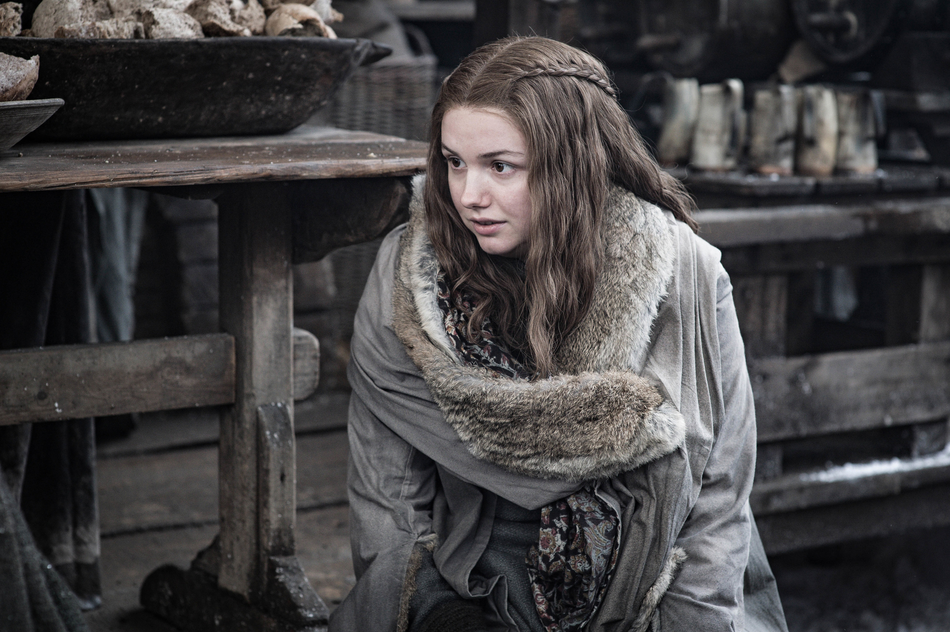 A young woman with braided brown hair and a fur-lined coat sits in a marketplace.