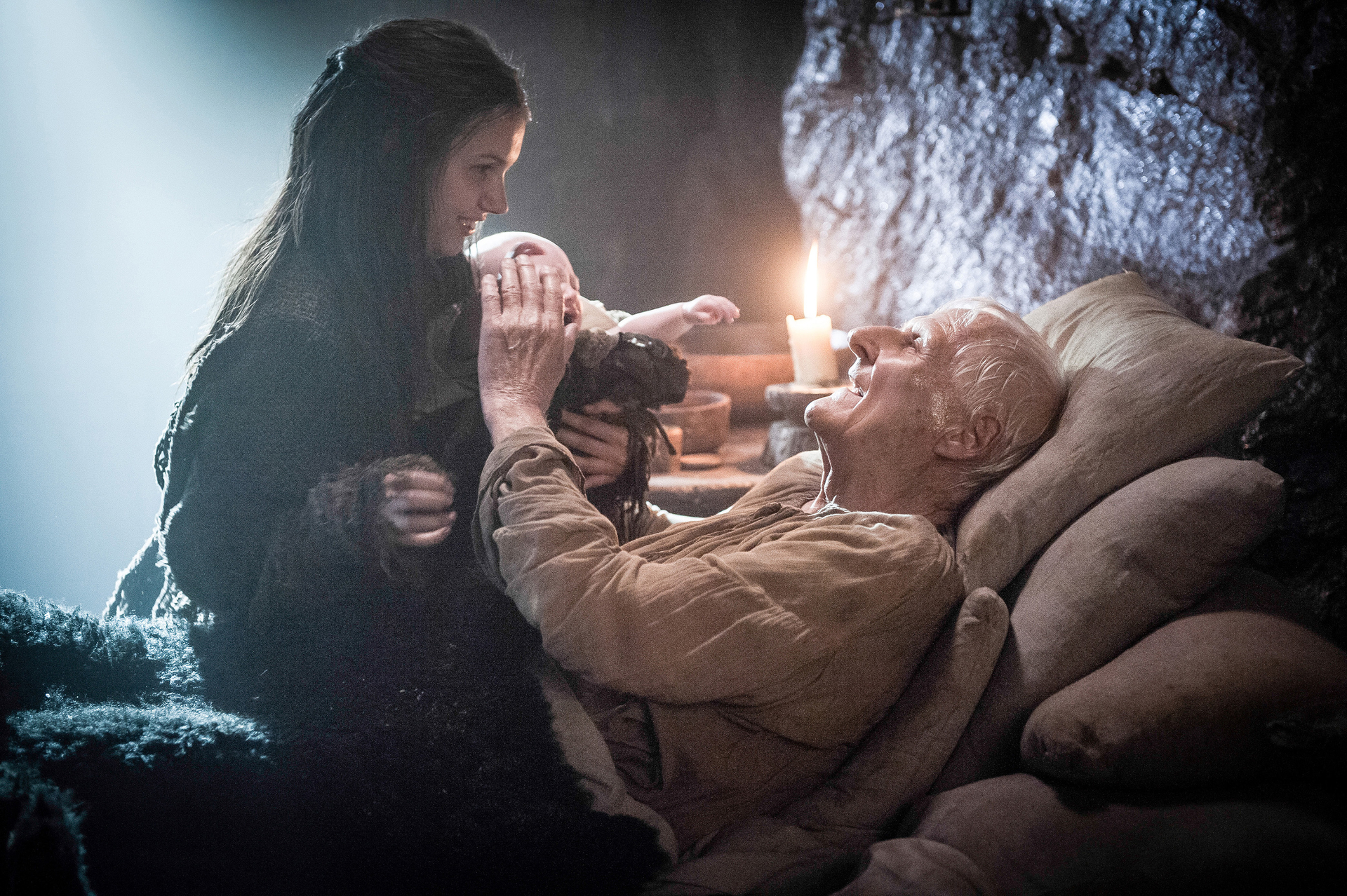 A woman holding a baby smiles at an old man lying in bed, who also smiles up at the baby.