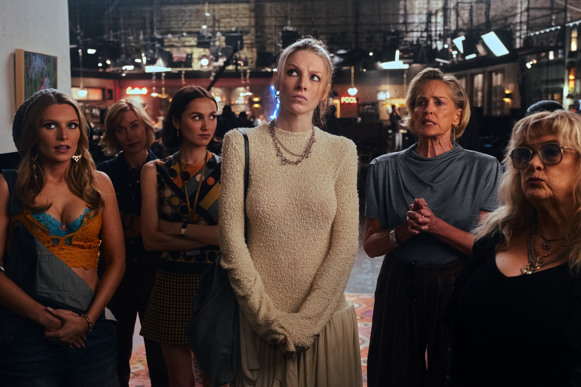 Five women in a varied outfits look forward with expressions ranging from concern to disinterest.