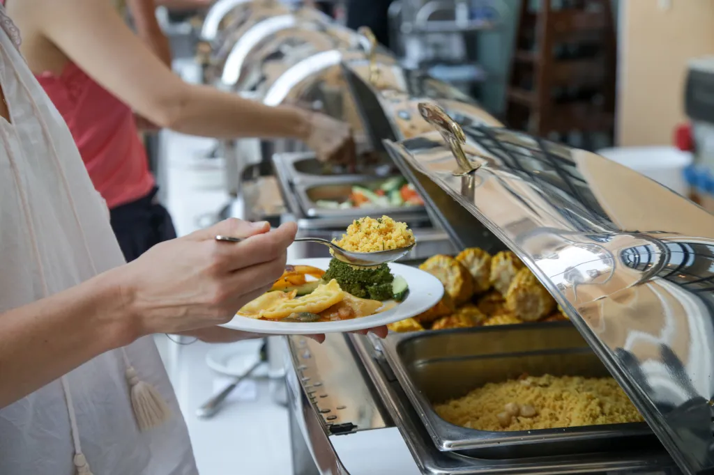 A woman serving herself food from a buffet line, including ravioli, broccoli, and couscous.
