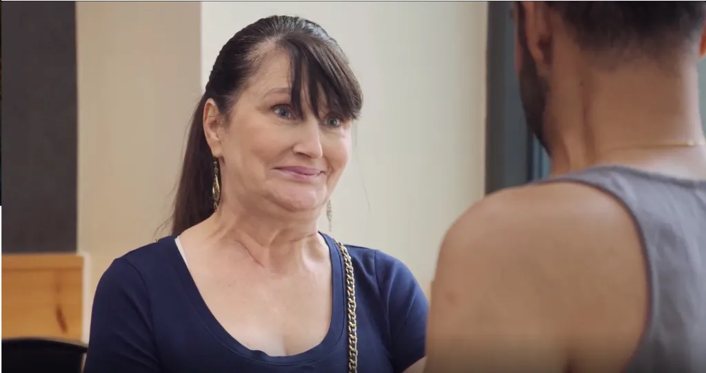 A woman in a blue shirt with a gold chain strap looks at a man wearing a grey tank top whose back is to the camera.