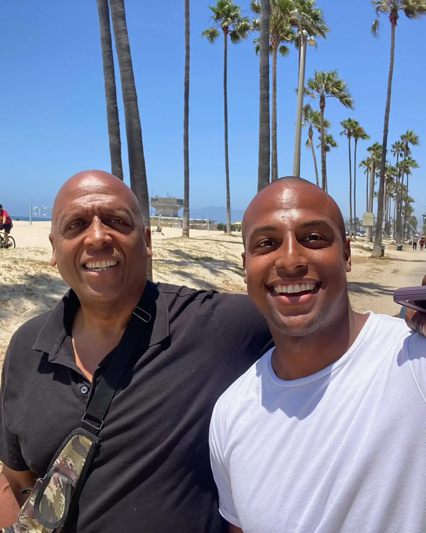 Two smiling bald men on a beach with palm trees.