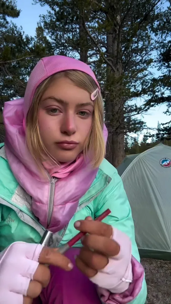 A person with blonde hair in a pink hood and mint green jacket holds a lipstick, with tents and trees in the background.