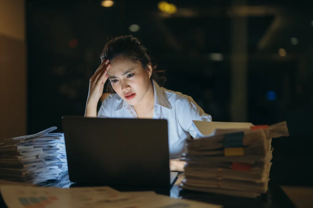A stressed woman with a headache looks at her laptop surrounded by stacks of paper in a dark office at night.