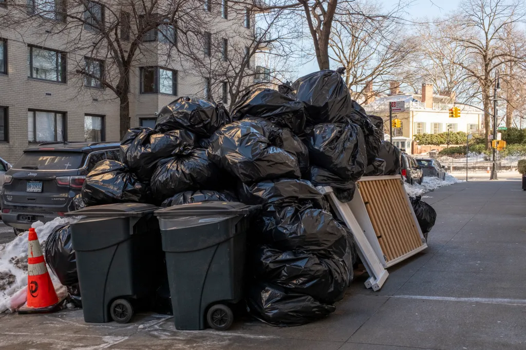 A large pile of black trash bags next to two gray trash cans and a wooden gate on a snowy street in New York City.