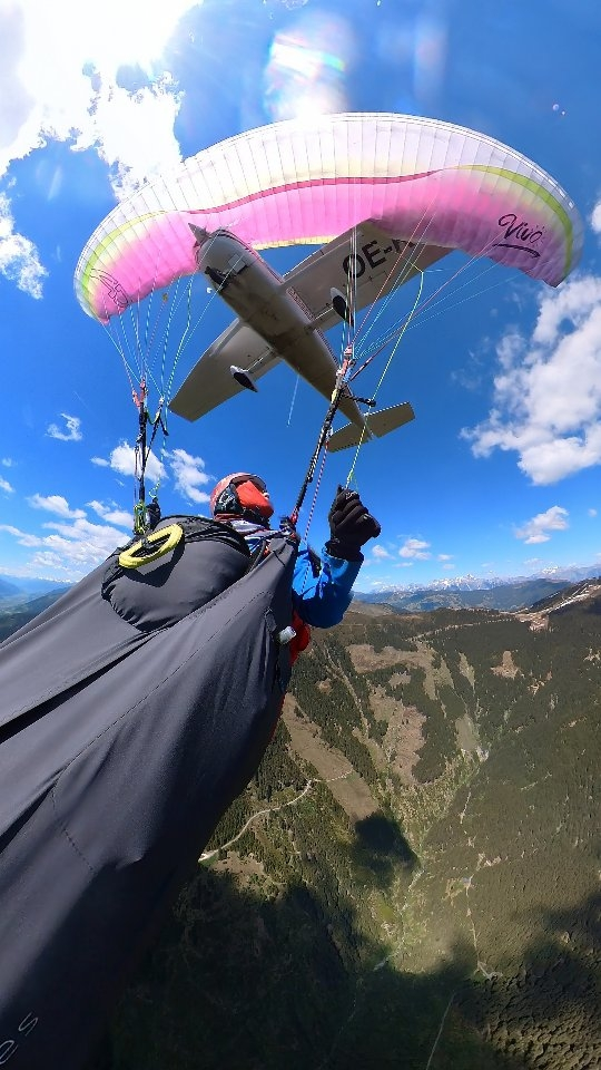 A paraglider, with a pink and white canopy, flying beneath a Cessna 172 plane over a mountainous landscape near Zell am See, Austria.
