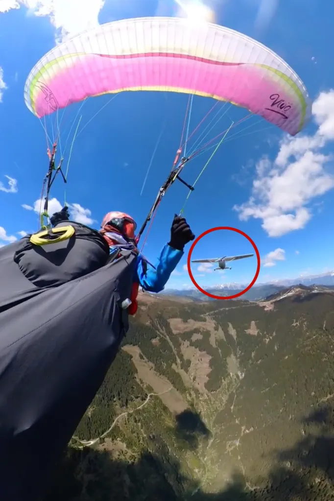 A paraglider soaring over mountains with a small plane circled in red in the distance.
