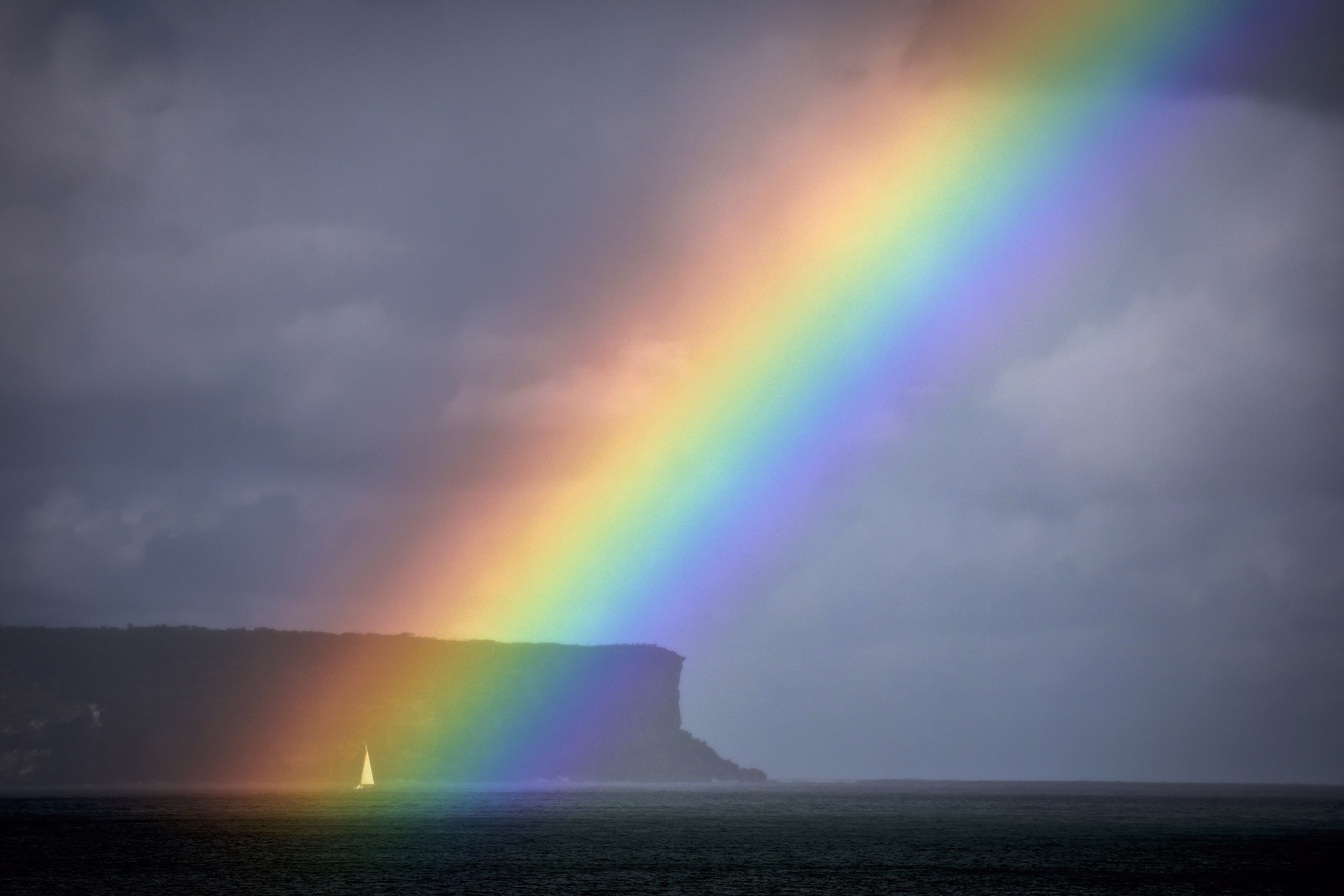 A sailboat passes a rainbow in front of a coastal cliff.