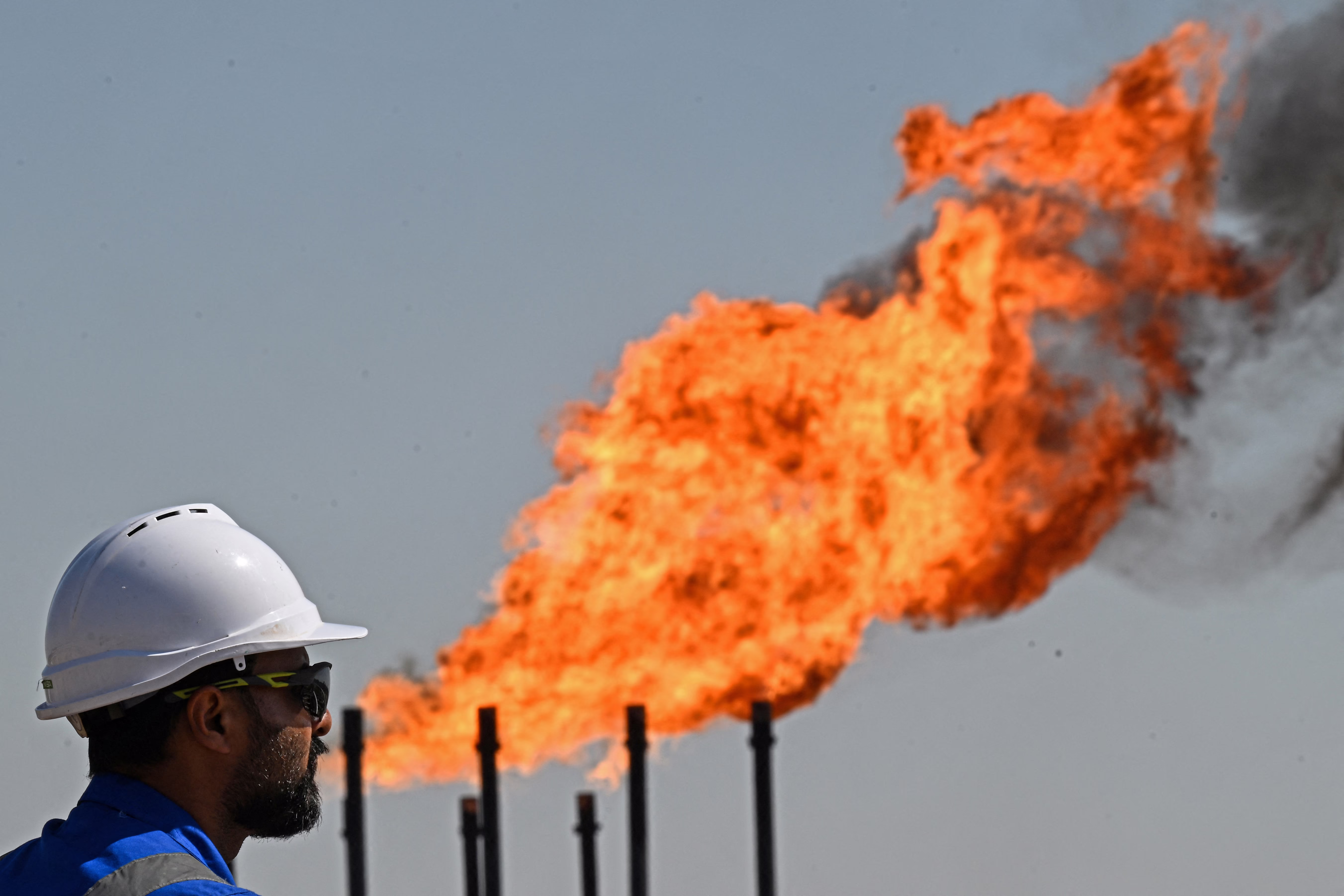 An oil company worker stands near a burning flare of gas. The photo is framed so the worker almost appears to be blowing the fire out of their mouth.