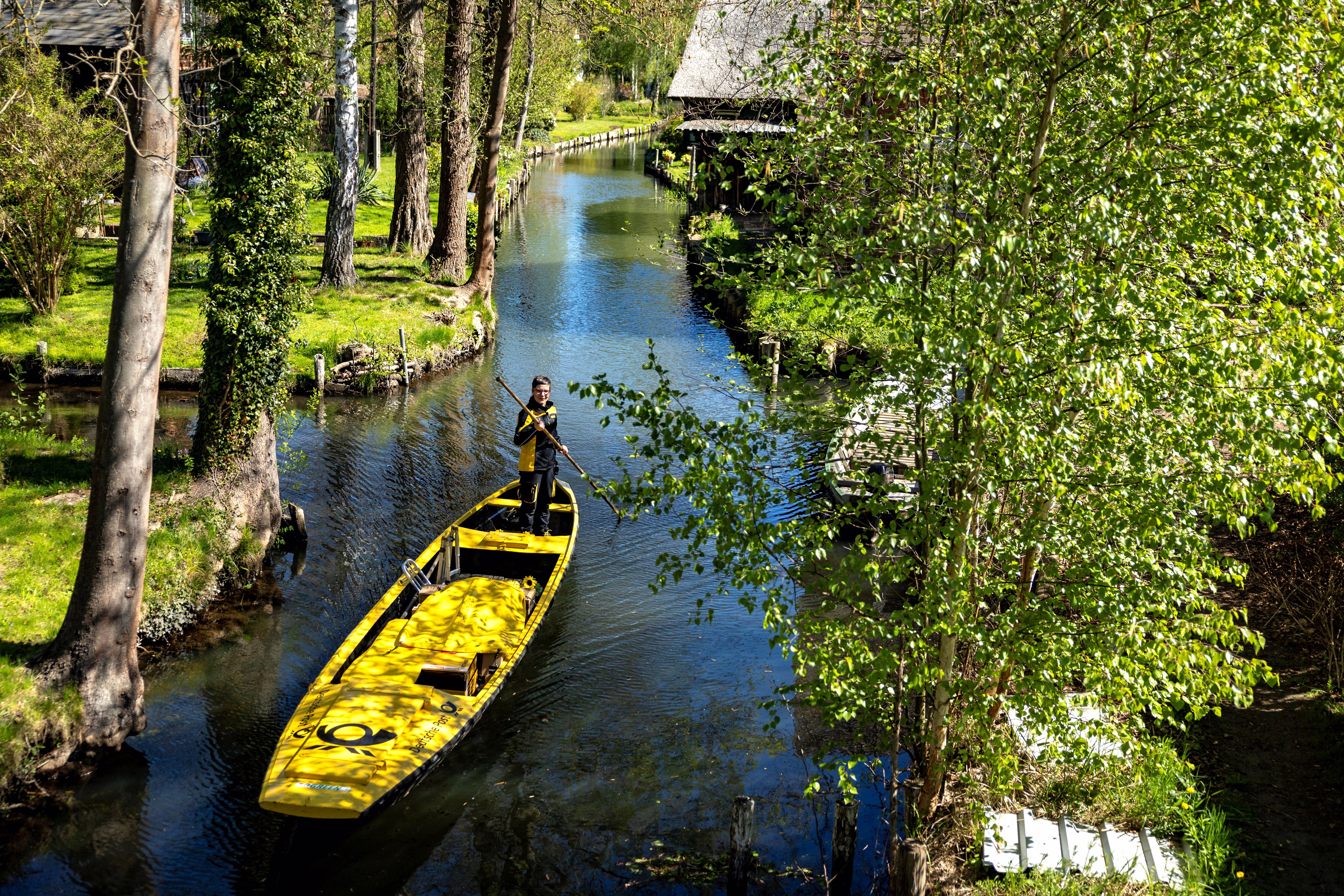 A person uses a pole to steer a small boat along a narrow canal through a forested neighborhood.