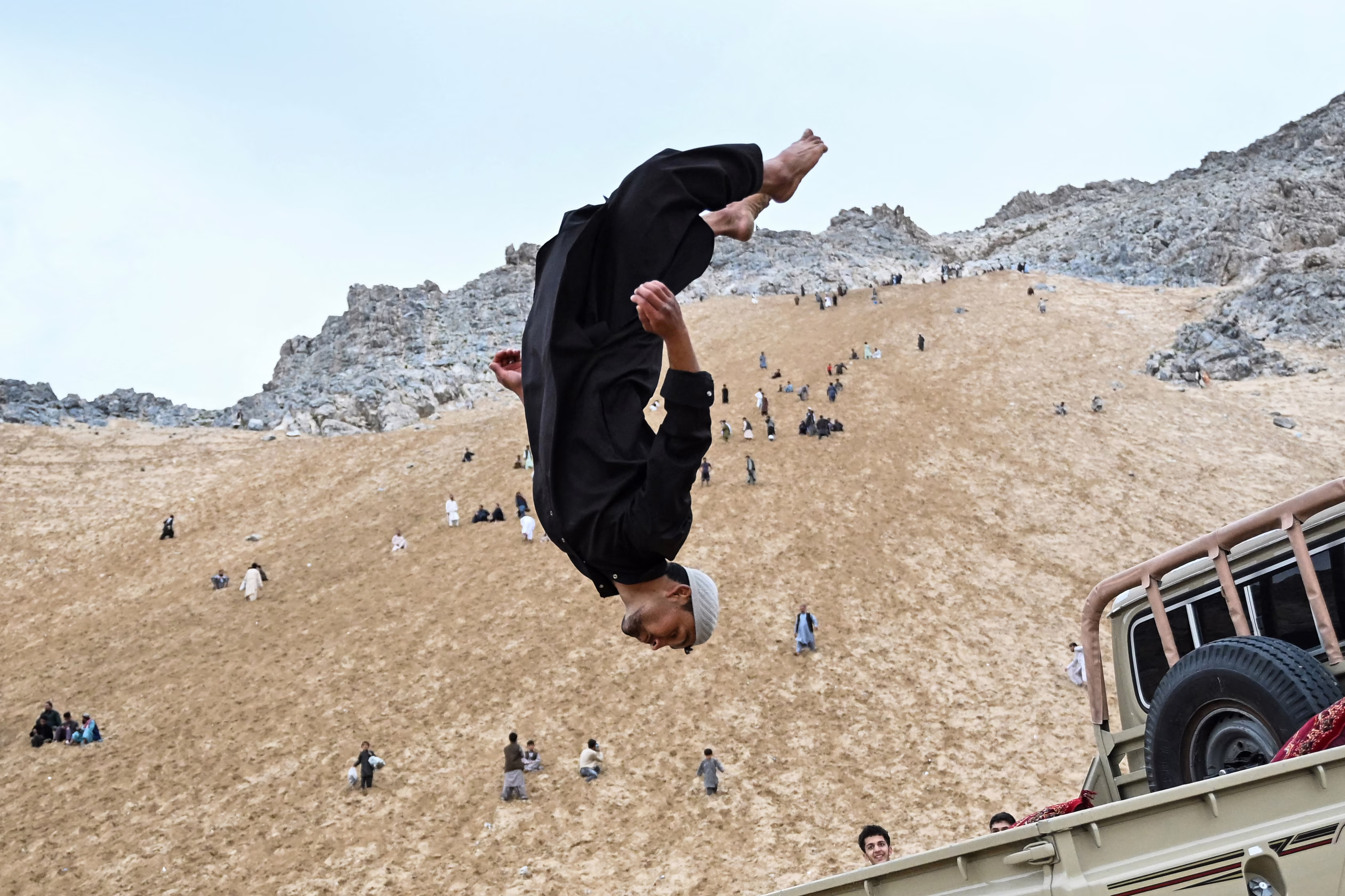 A parkour athlete leaps upside down, performing a stunt off the back of a vehicle on a sandy mountainside.