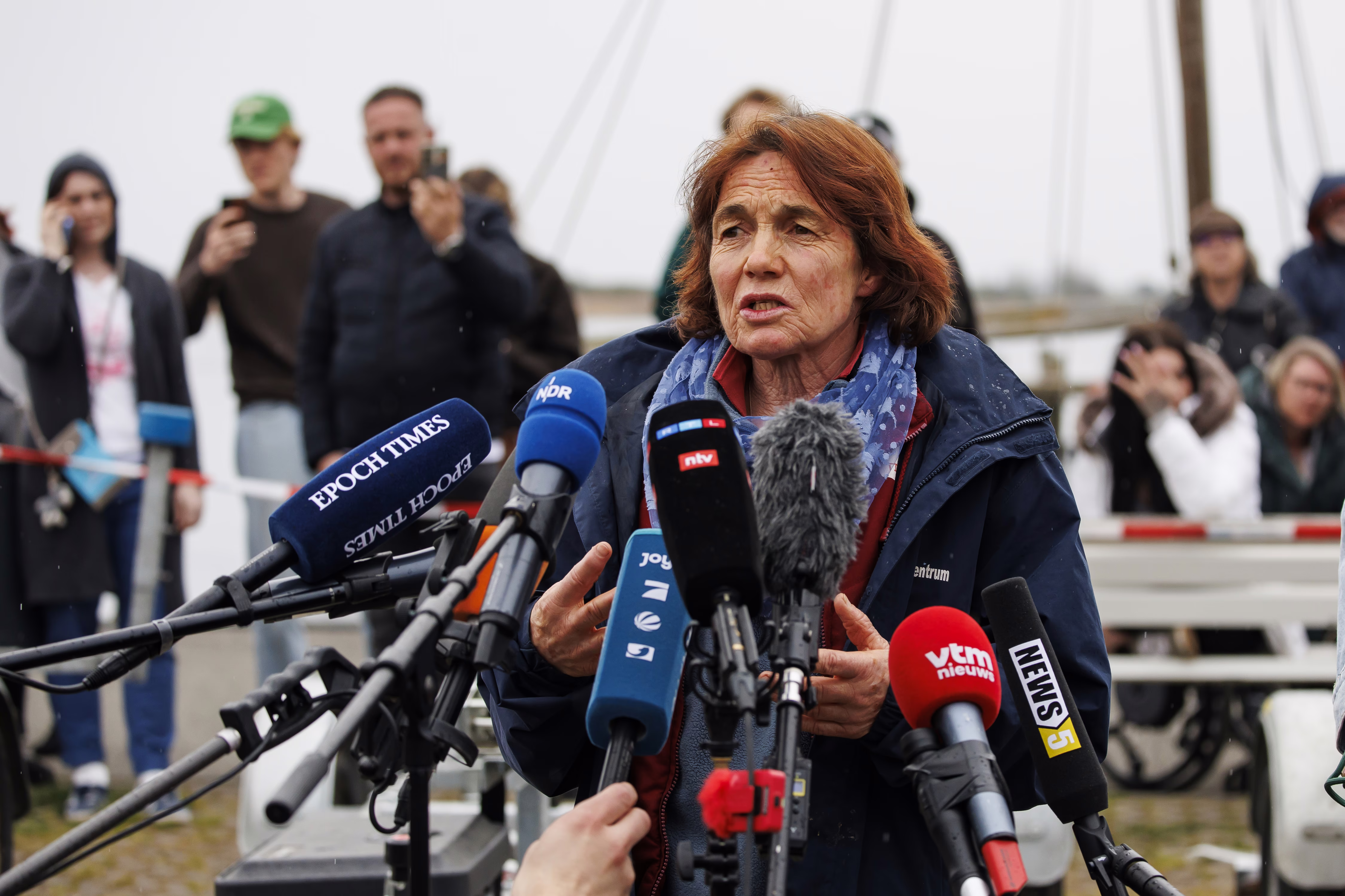 A person speaks to reporters, standing outside in front of a 8 or 9 microphones.