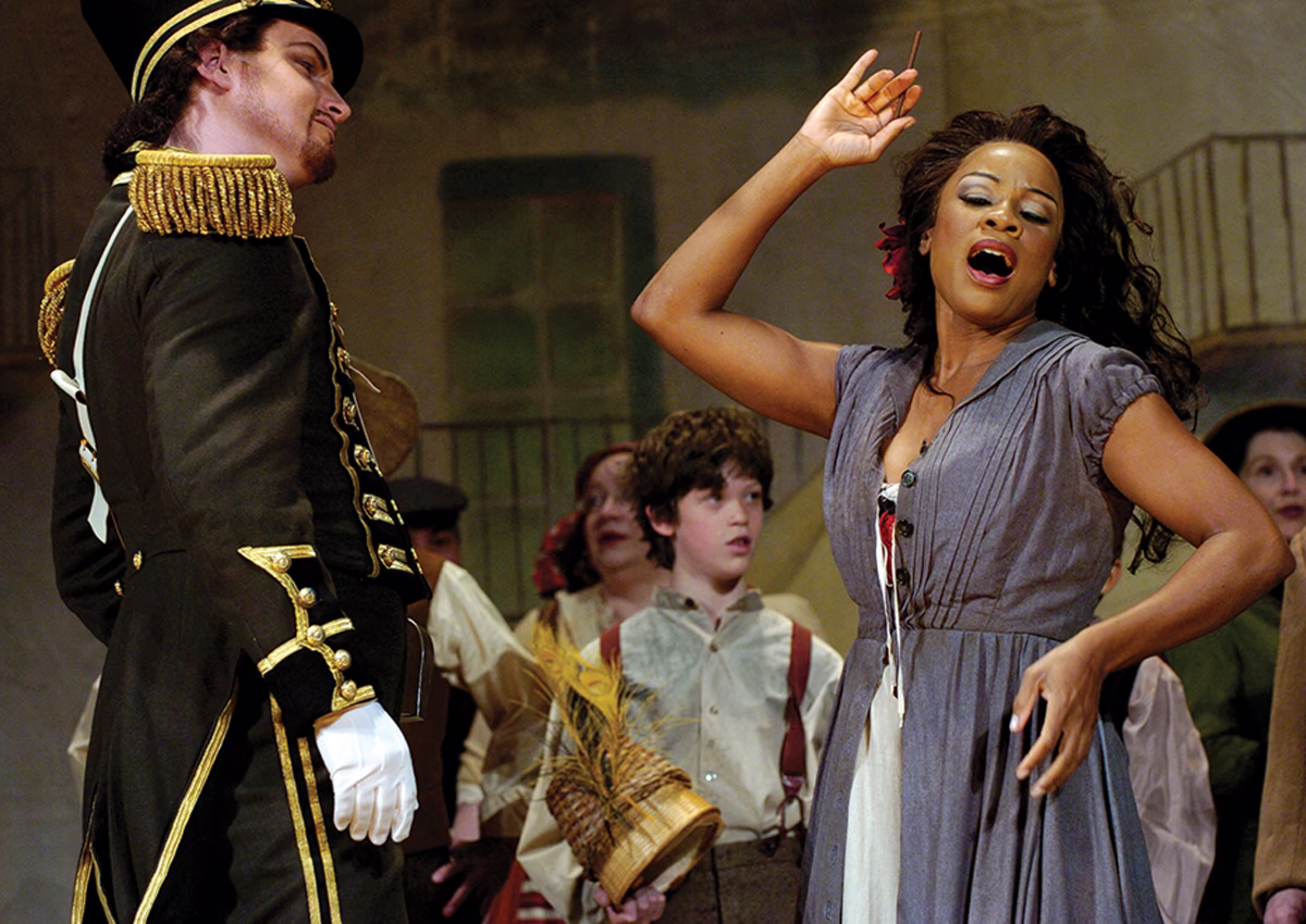 photo of woman on opera stage singing with arm raised dramatically, with man in uniform listening and children in background