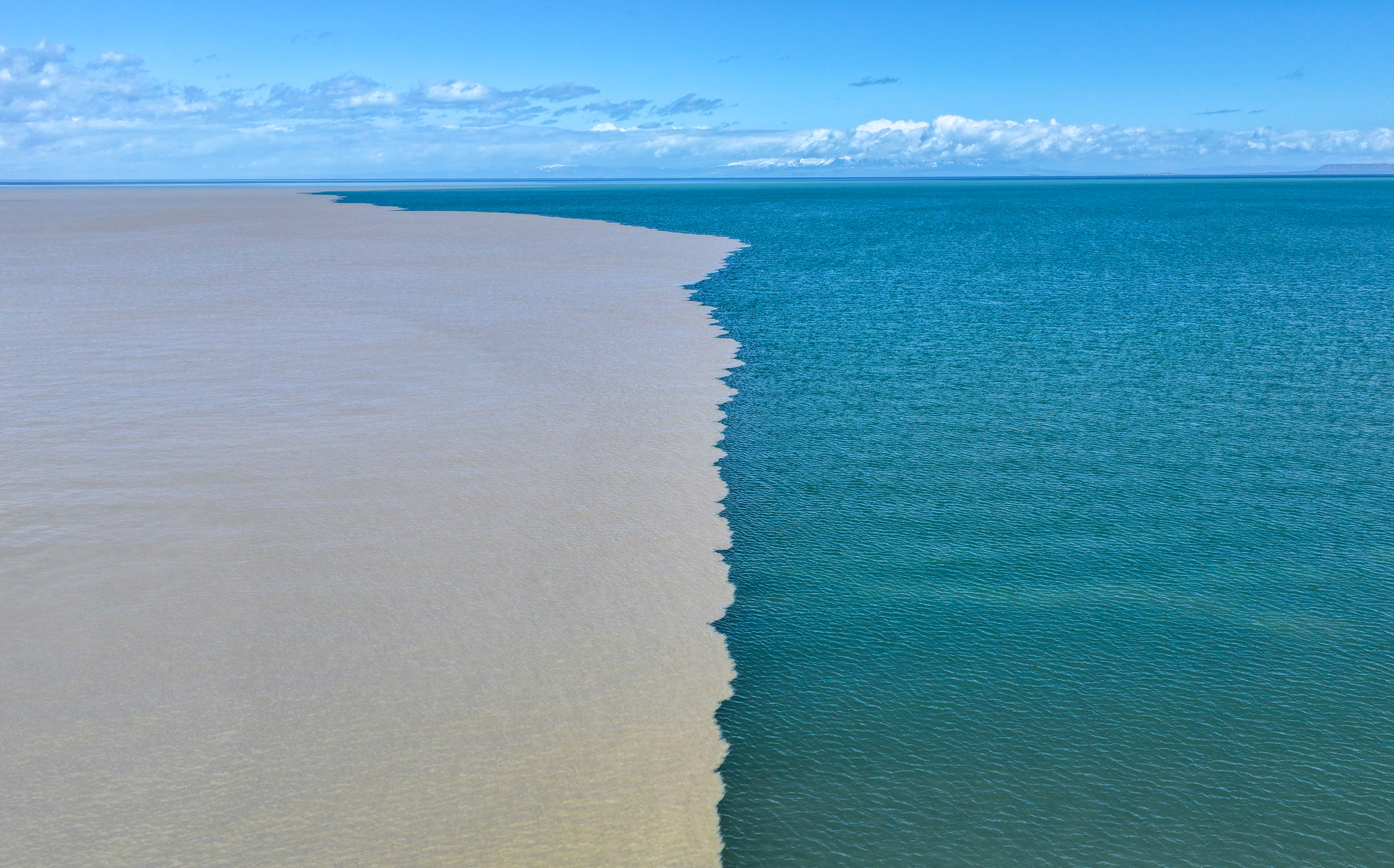 An aerial view of an expanse of lake water, roughly split in half with the left side being muddy water and the right side being clear blue water.