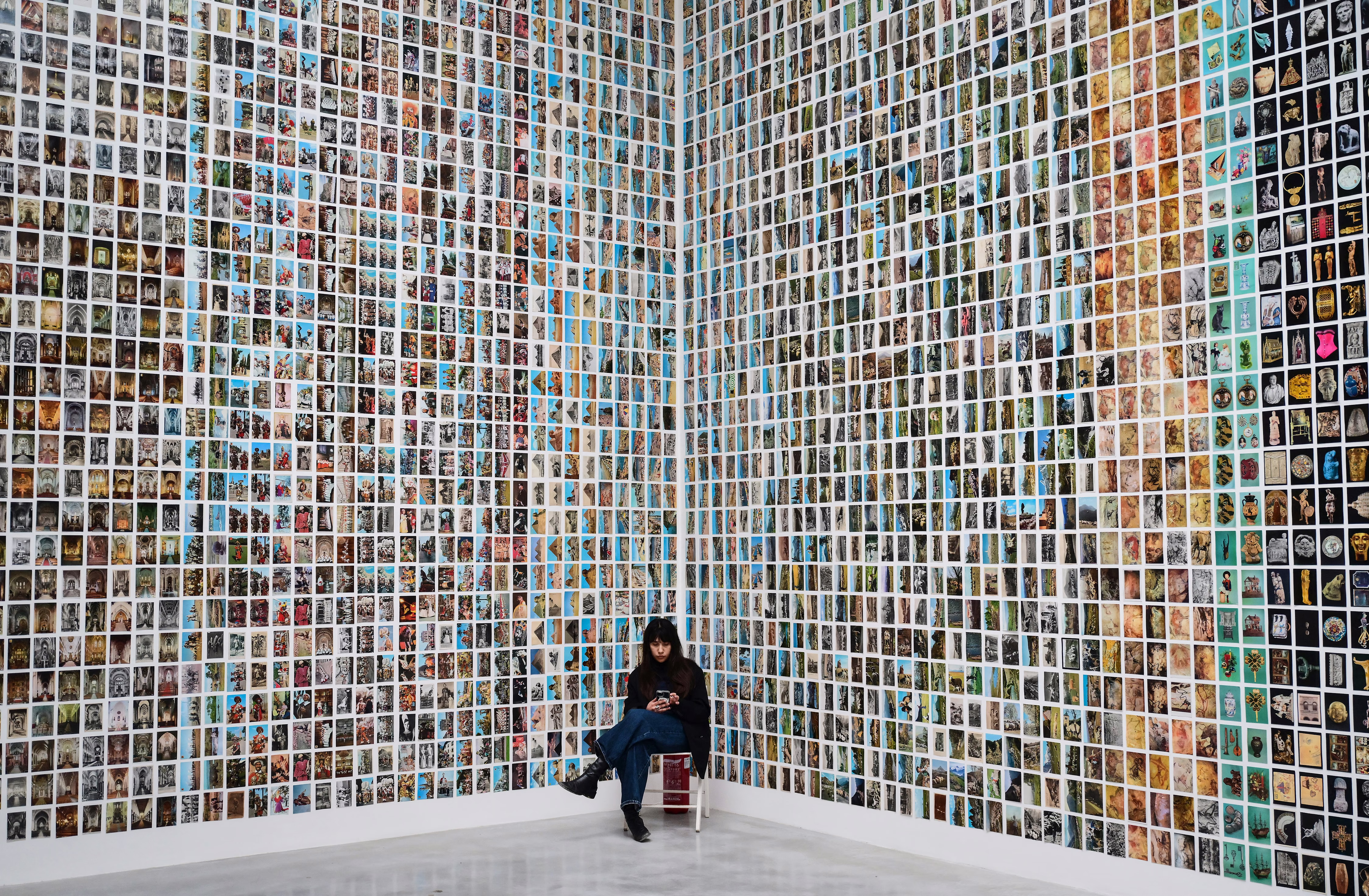 A gallery attendant sits in a corner beside two tall walls covered in hundreds of photographs. 