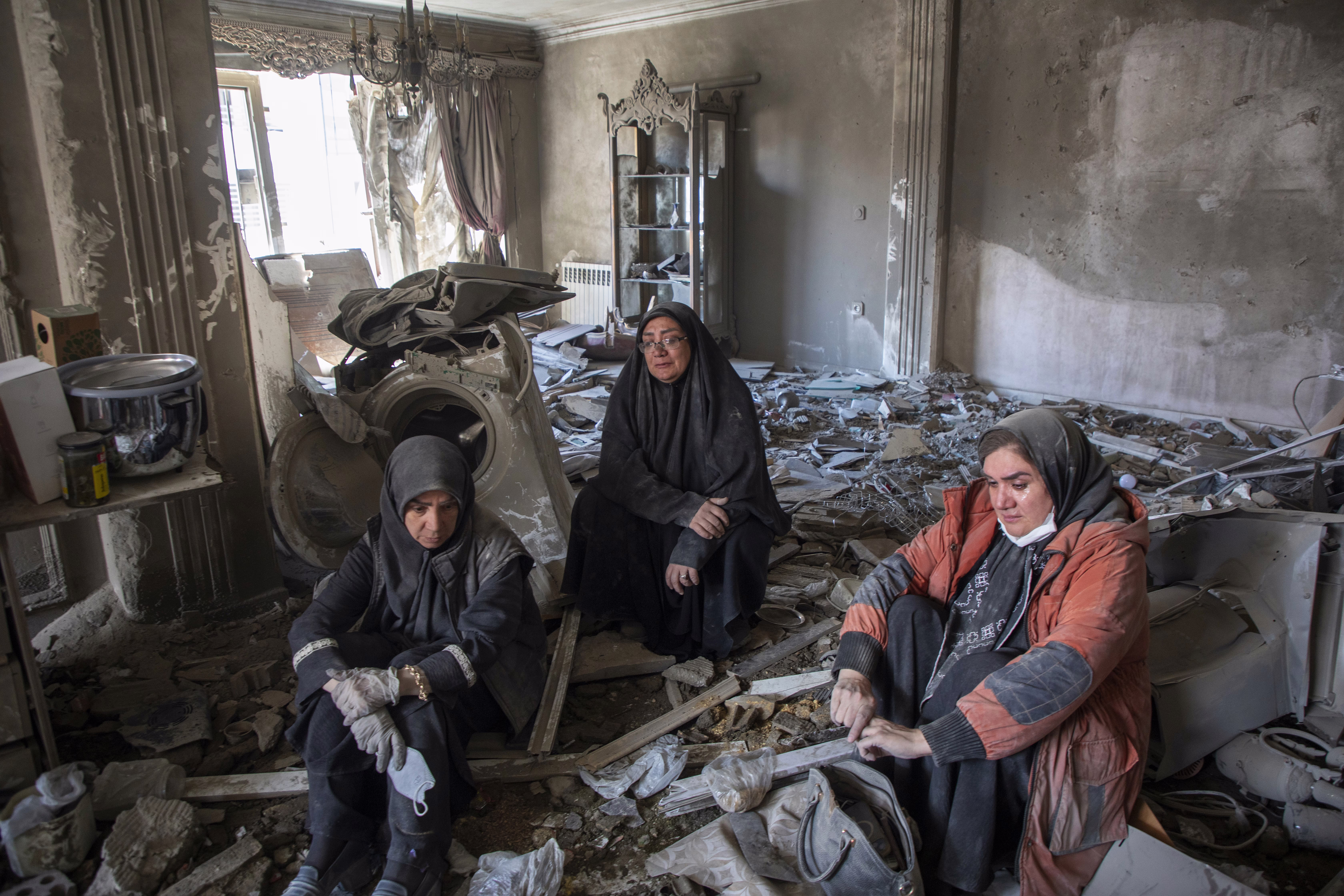 Three people sit inside a heavily-damaged room, among dust and debris, after an airstrike.