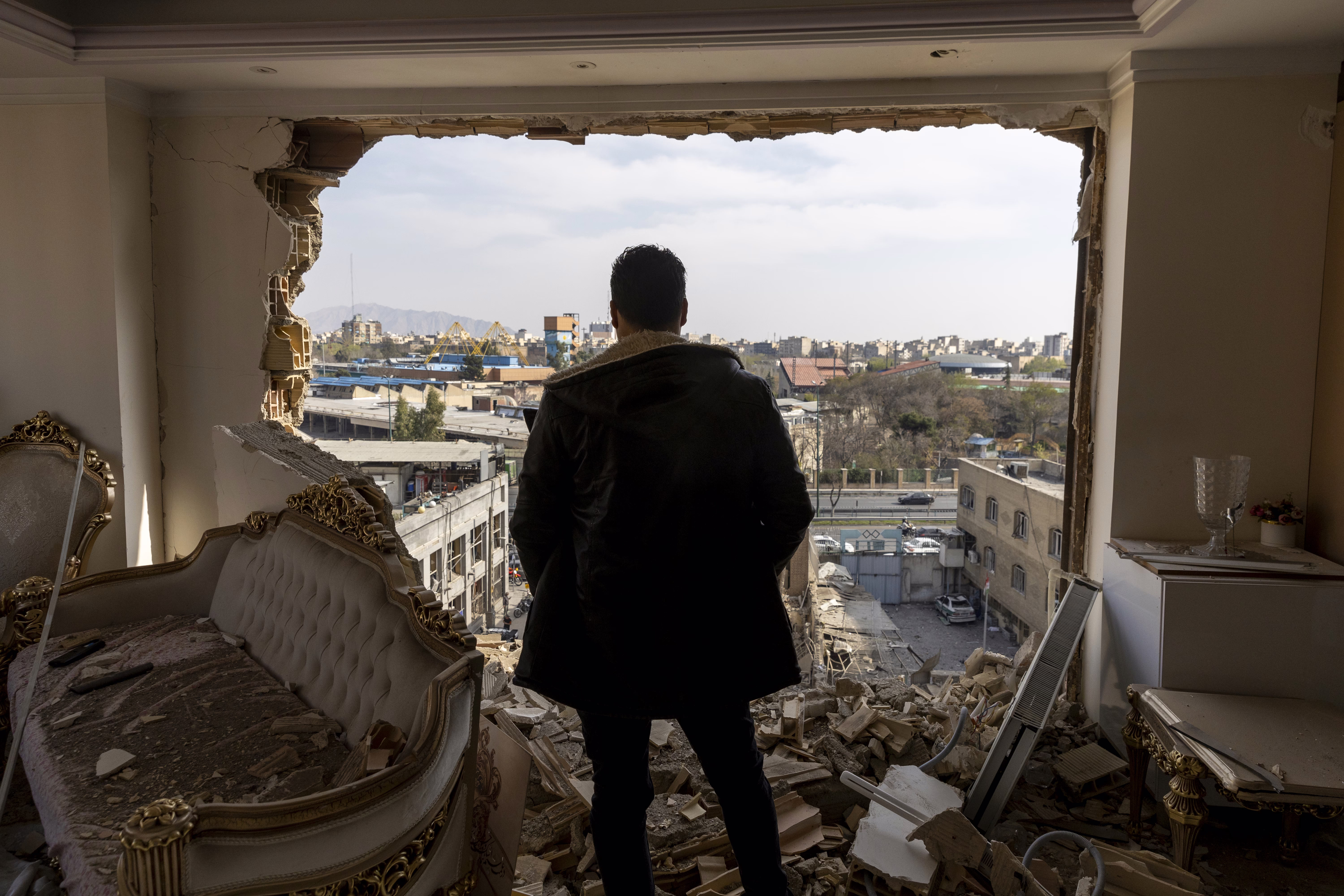 A man stands in a damaged residence, looking out of a blown-out wall, toward other buildings.