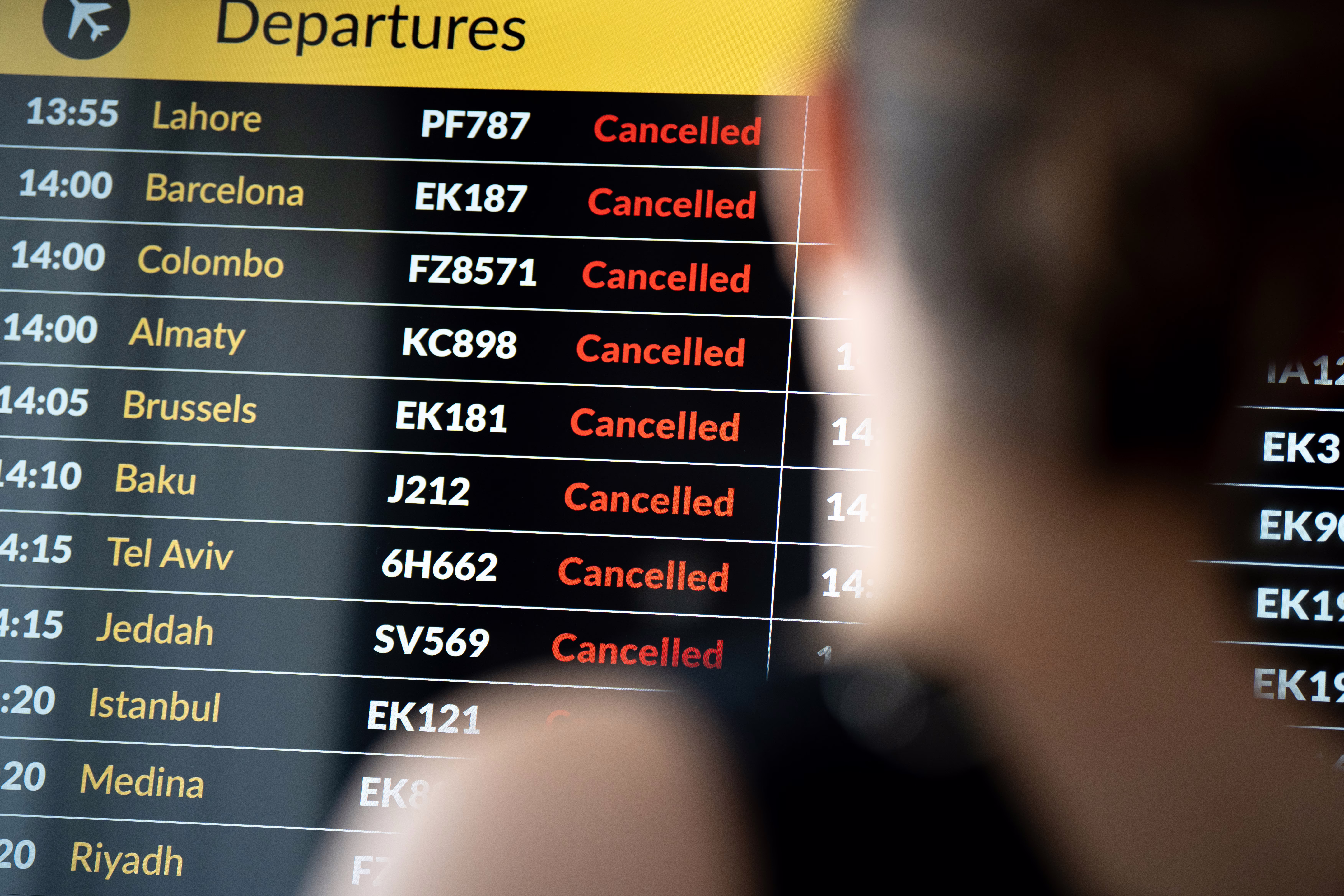 A passenger checks her flight status on an airport arrivals and departures board listing many cancellations.