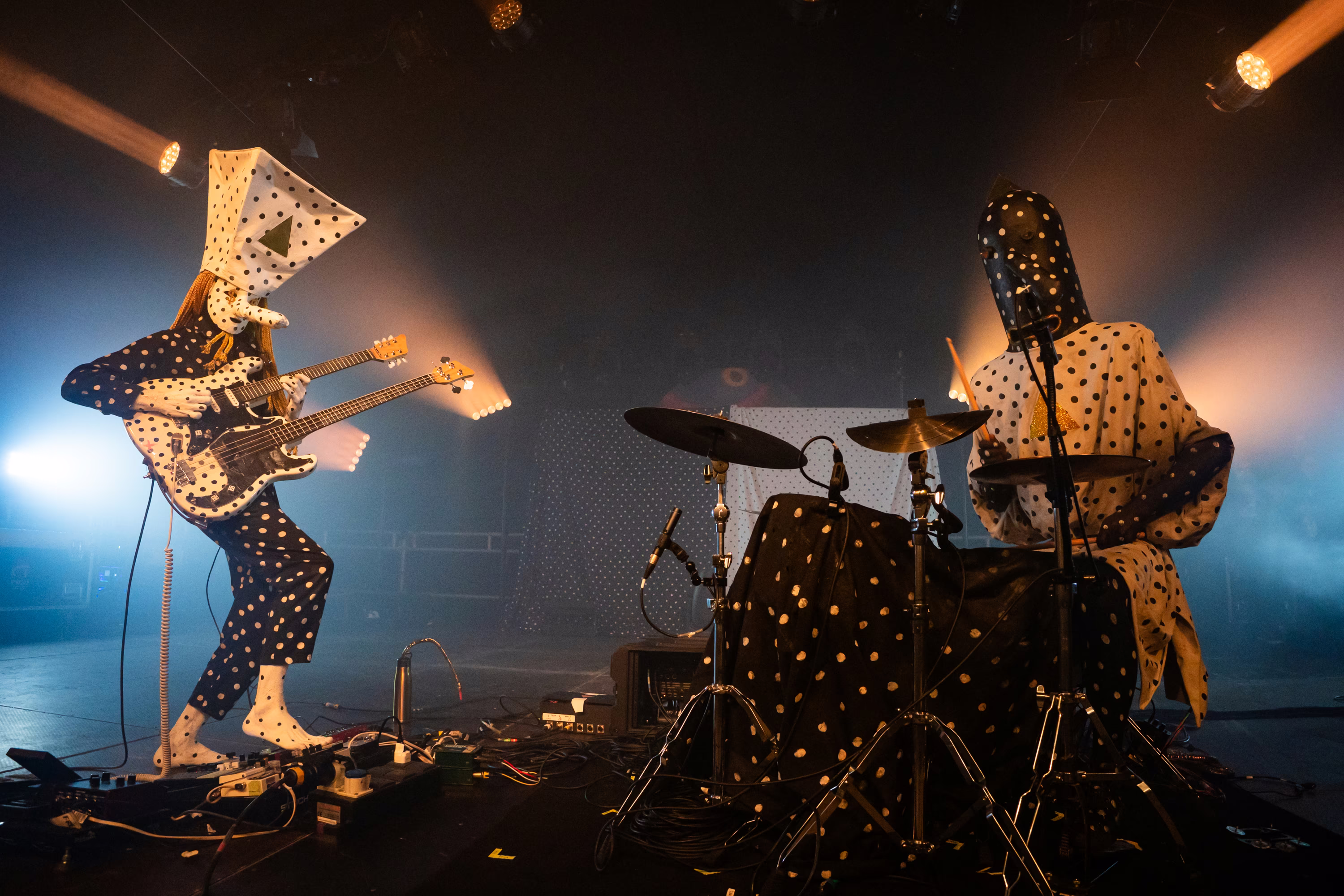 A two-person band performs on stage. The guitarist and drummer are both dressed in polkadot costumes and masks.