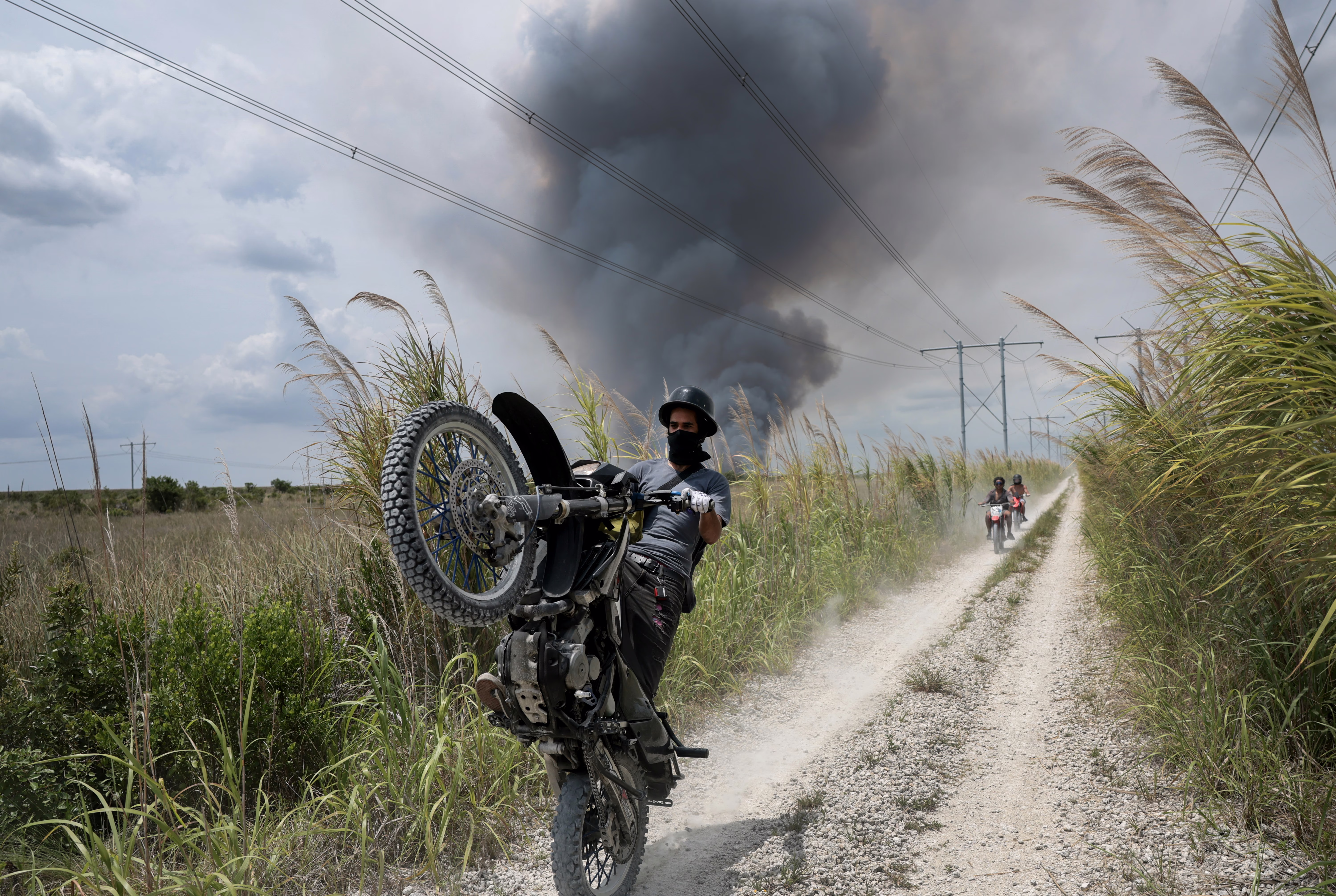 A motorcycle rider pops a wheelie in front of the photographer, while riding on a dirt road through grassland, with dark smoke rising in the background from a brushfire.