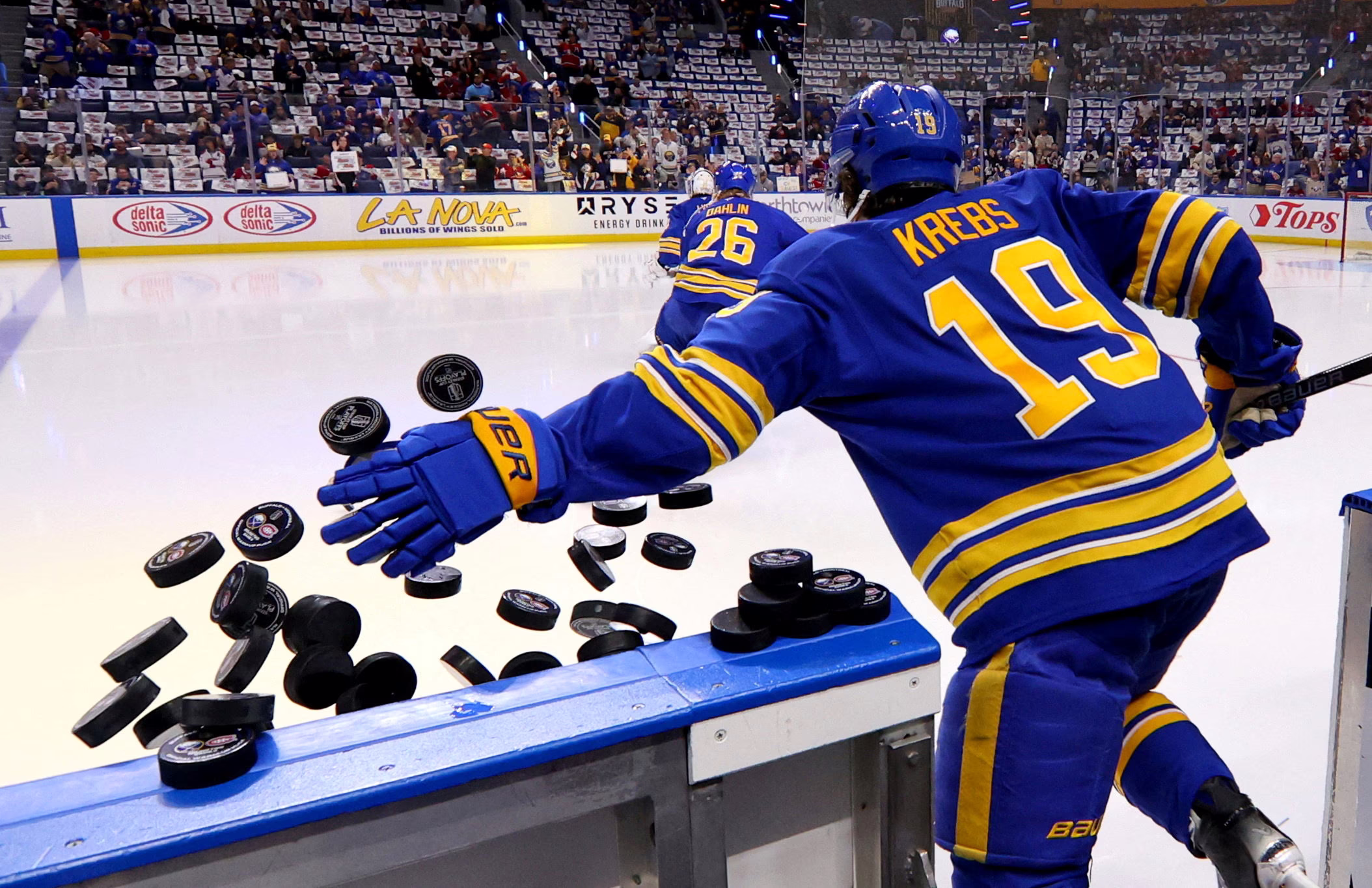 Professional hockey players enter an ice arena, knocking a pile of pucks off of a wall on the way.