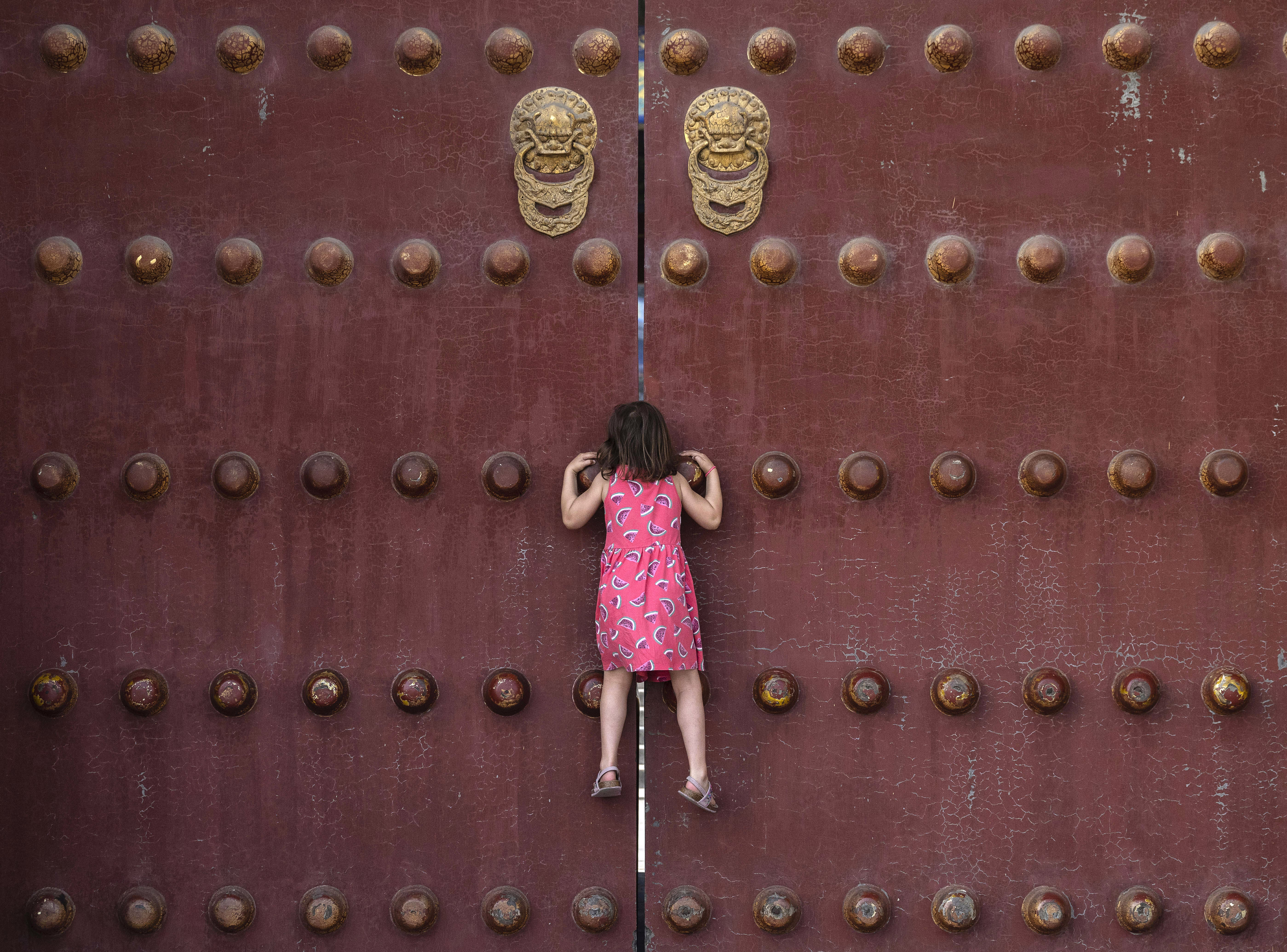 A young girl climbs to look through a gap of the large closed doors of a temple.