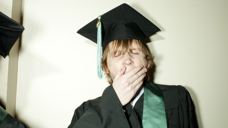 Photograph of a man wearing a cap and gown with a green sash over his shoulder covering his mouth as he yawns. His eyes are closed, as if sleepy.