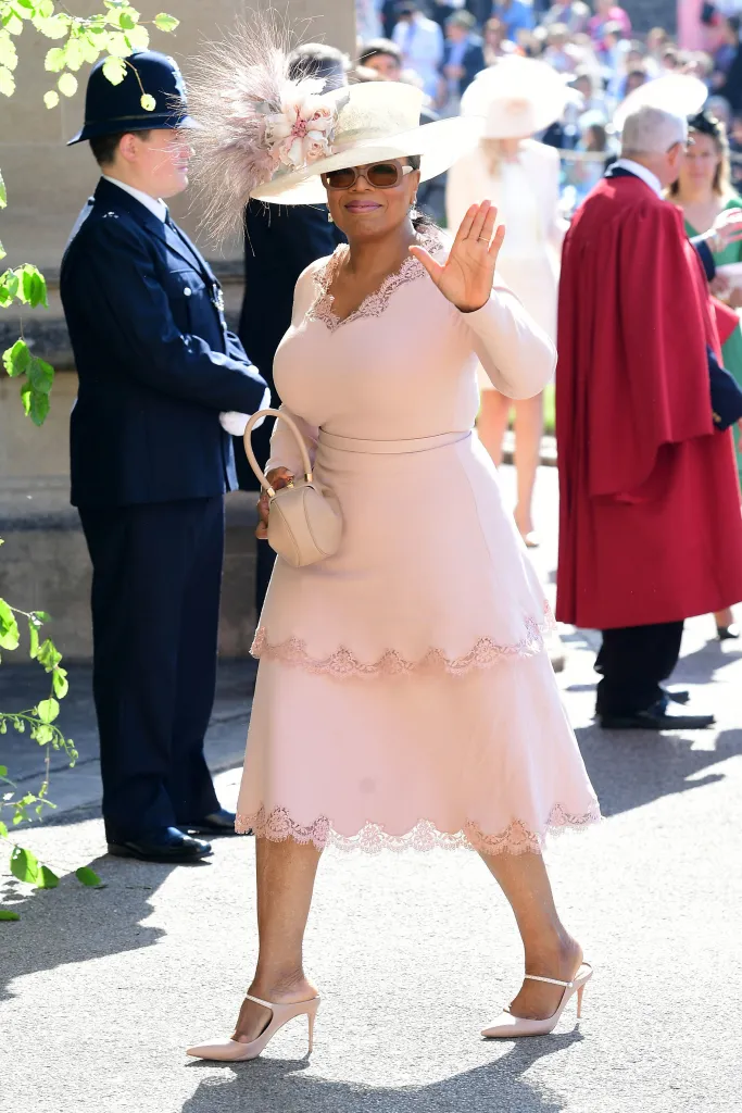 Oprah Winfrey in a pale pink dress and hat, waving to the camera.