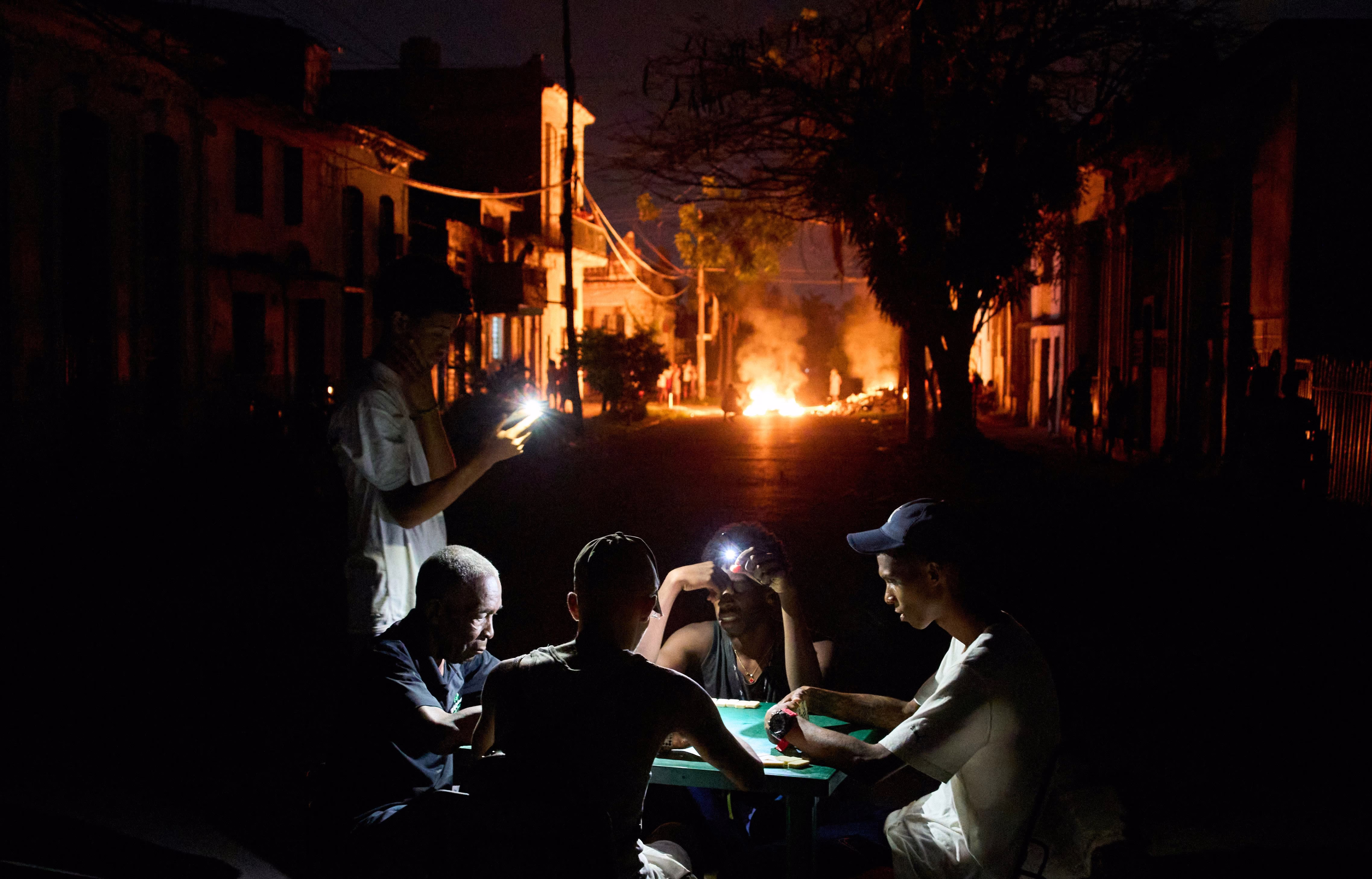 People illuminate themselves with their phones while playing dominoes; in the background, a fire, set up by residents protesting against prolonged power outages, burns on a street in Havana.