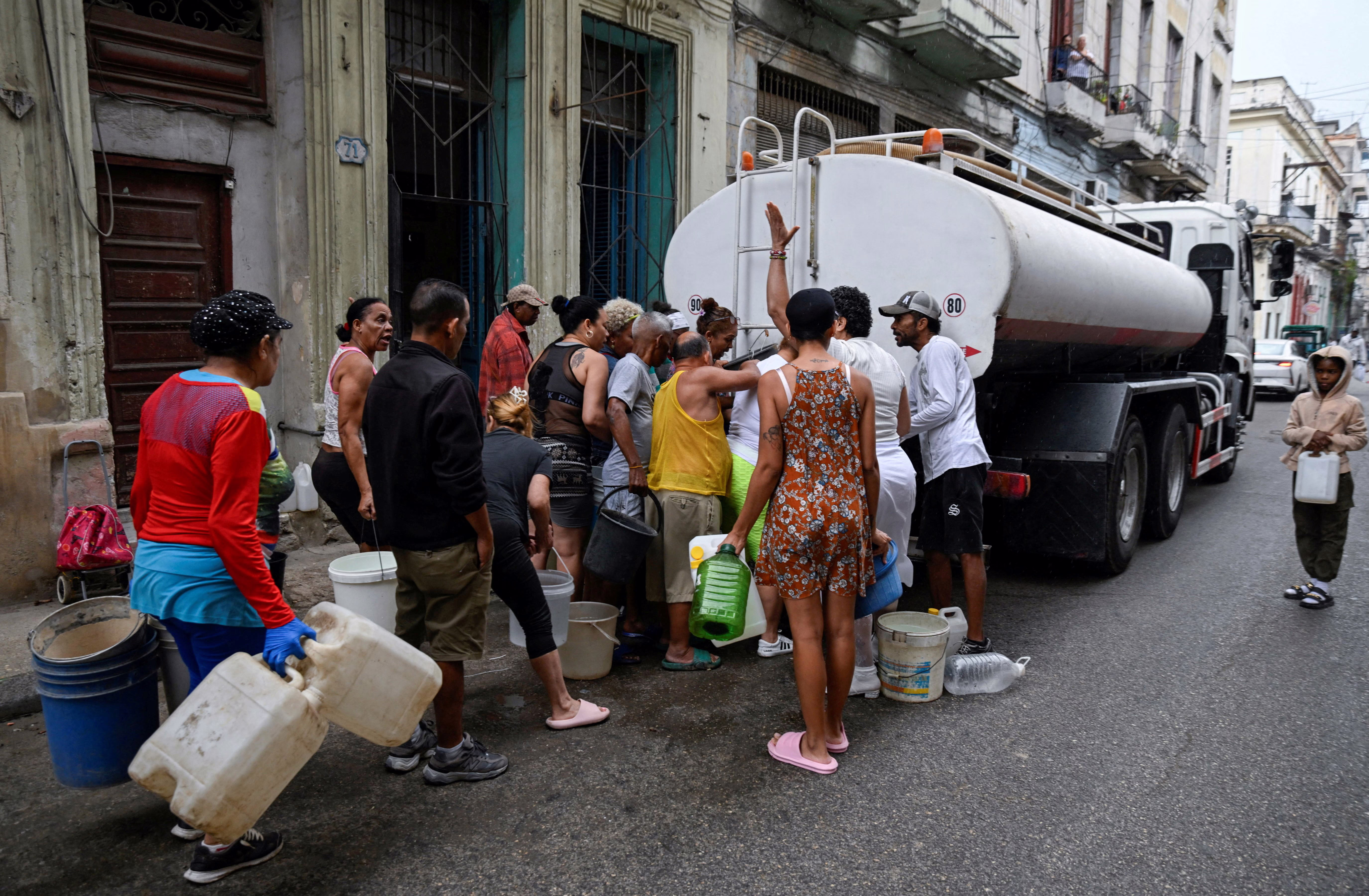 People gather around a water tanker truck to fill up buckets and other containers in a street.