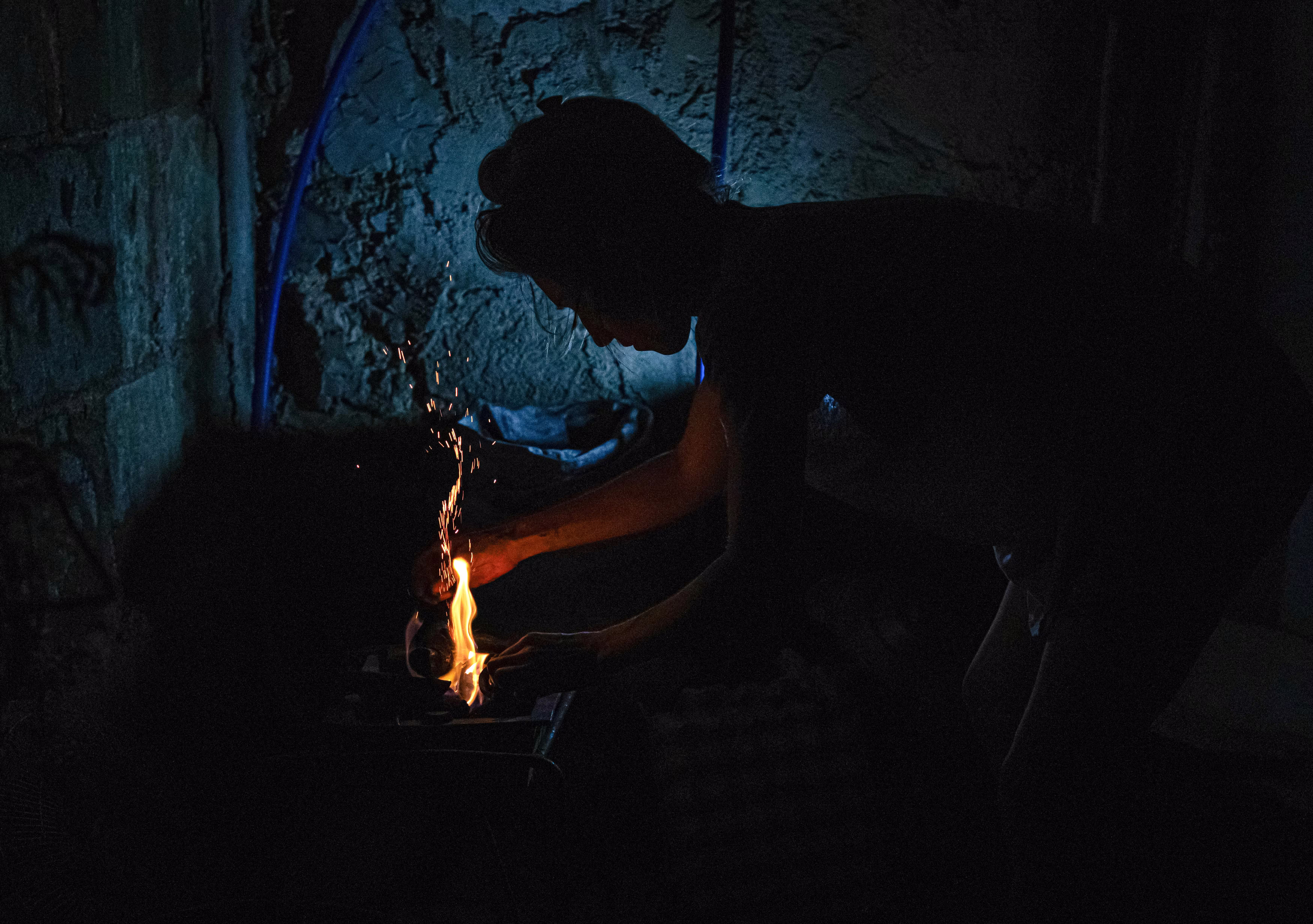 A woman prepares a wood fire to cook food inside a dark room.