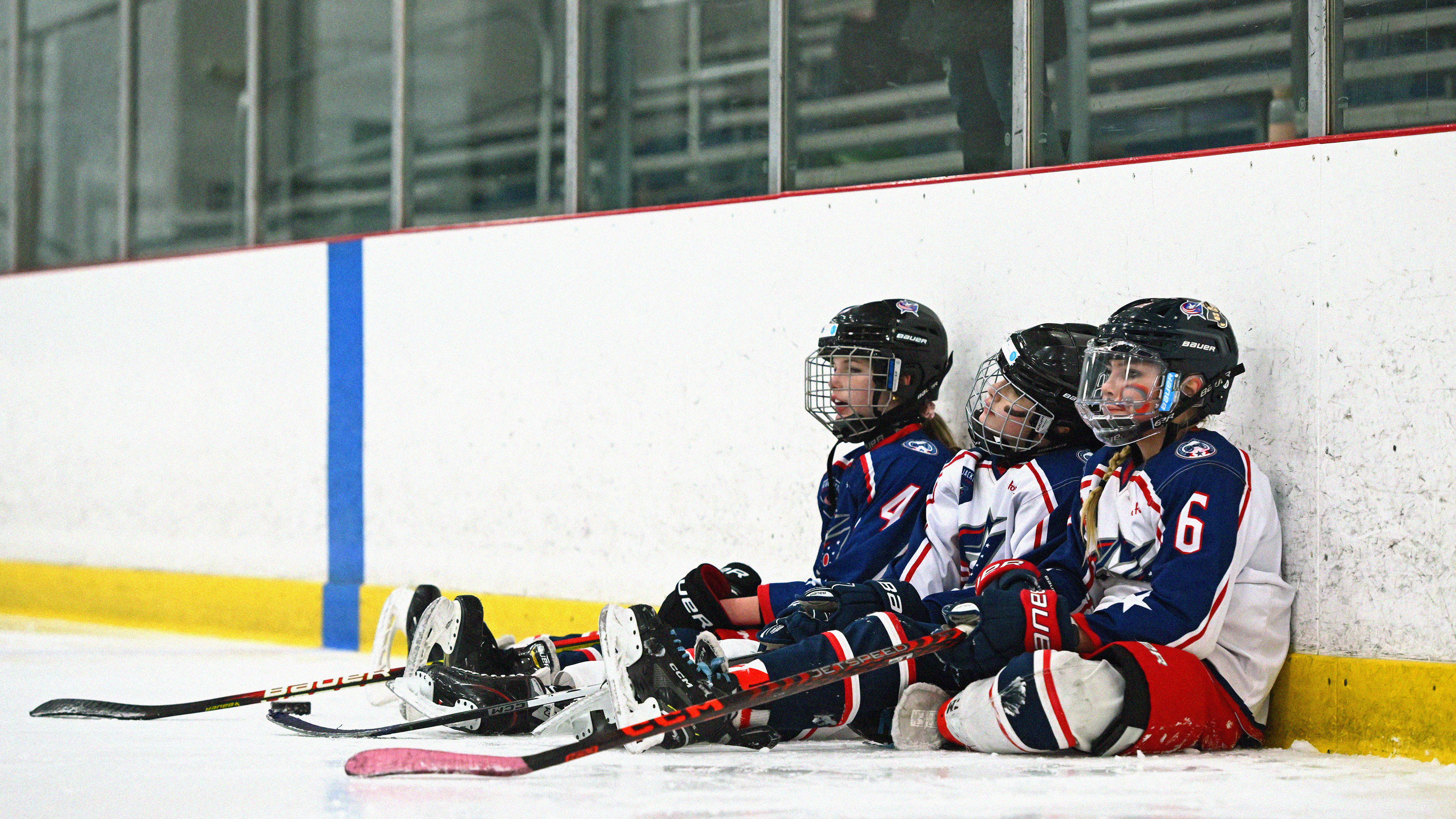 Photograph of three children in red, white, and blue hockey jerseys sitting down next to each other on the ice of a skating rink. They are holding hockey sticks and wearing helmets.