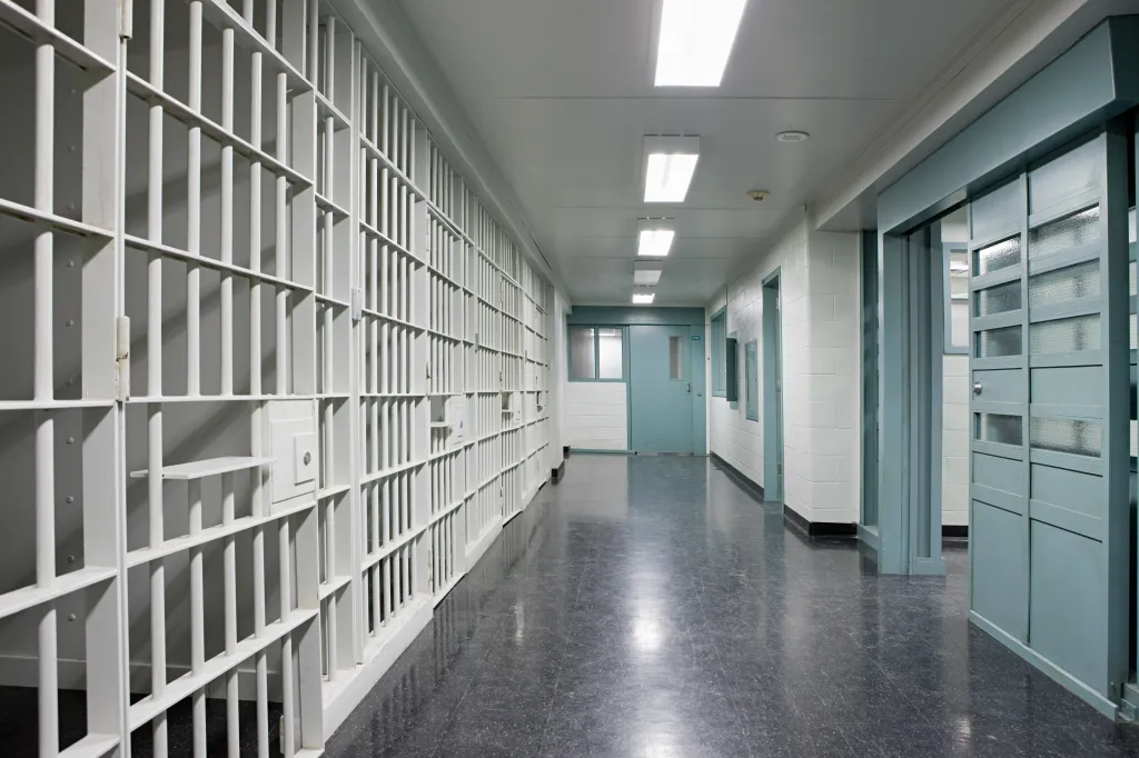 An empty prison corridor with white cell bars lining the left wall and teal-colored doors and frames on the right.