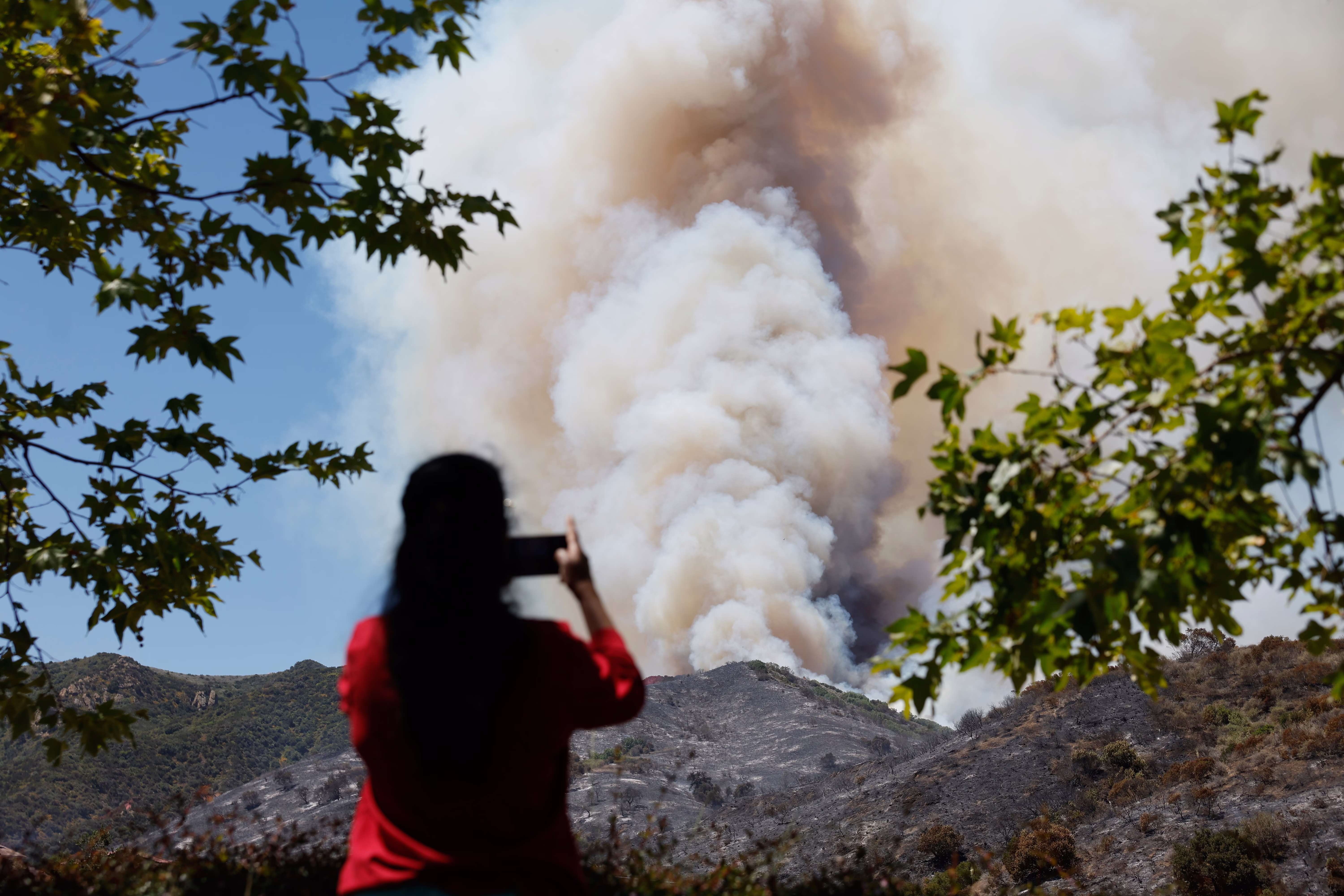 A person photographs a huge plume of smoke rising above a hillside wildfire.
