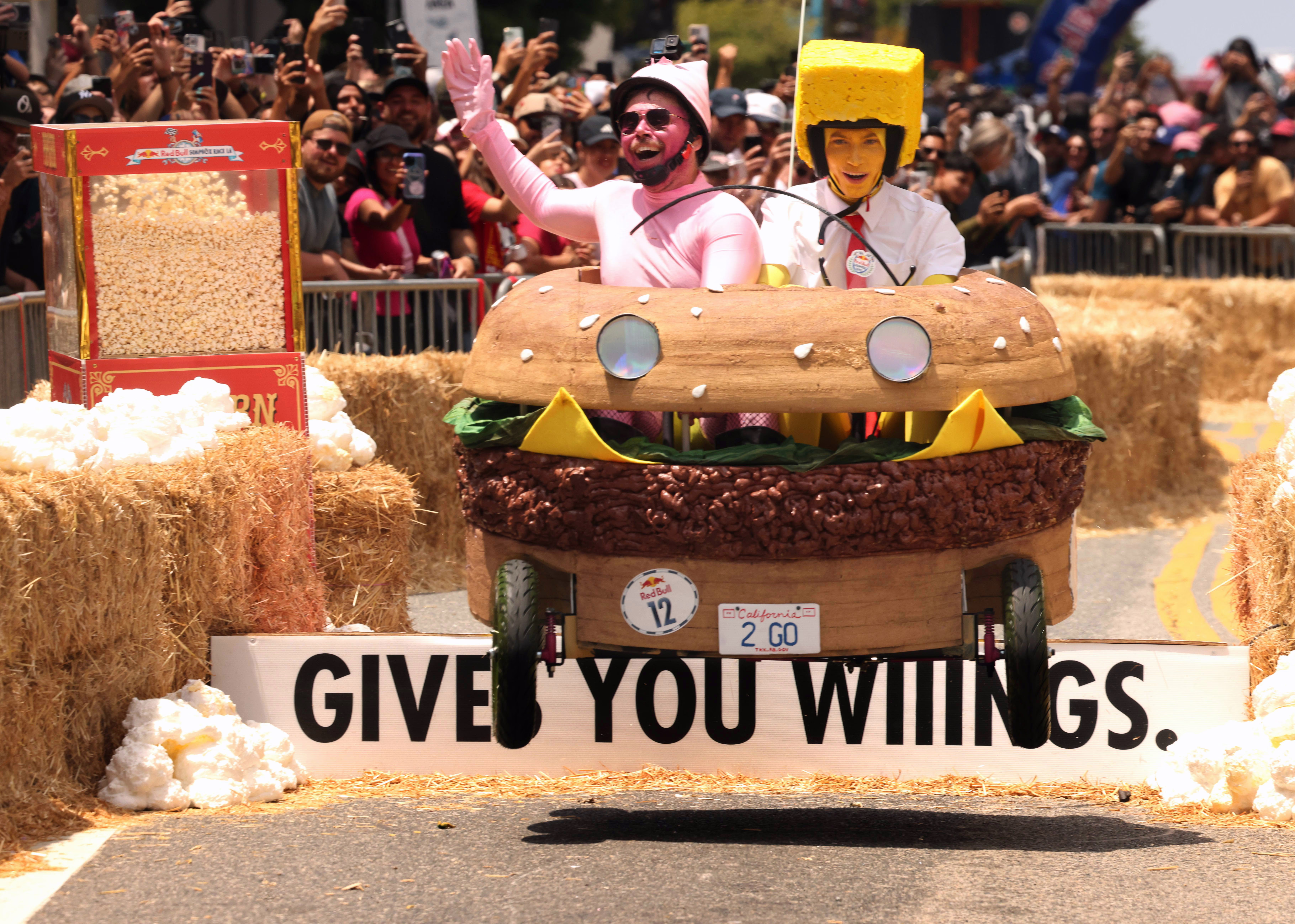 Two people in costumes race a hamburger-shaped soapbox-derby cart, making a jump.
