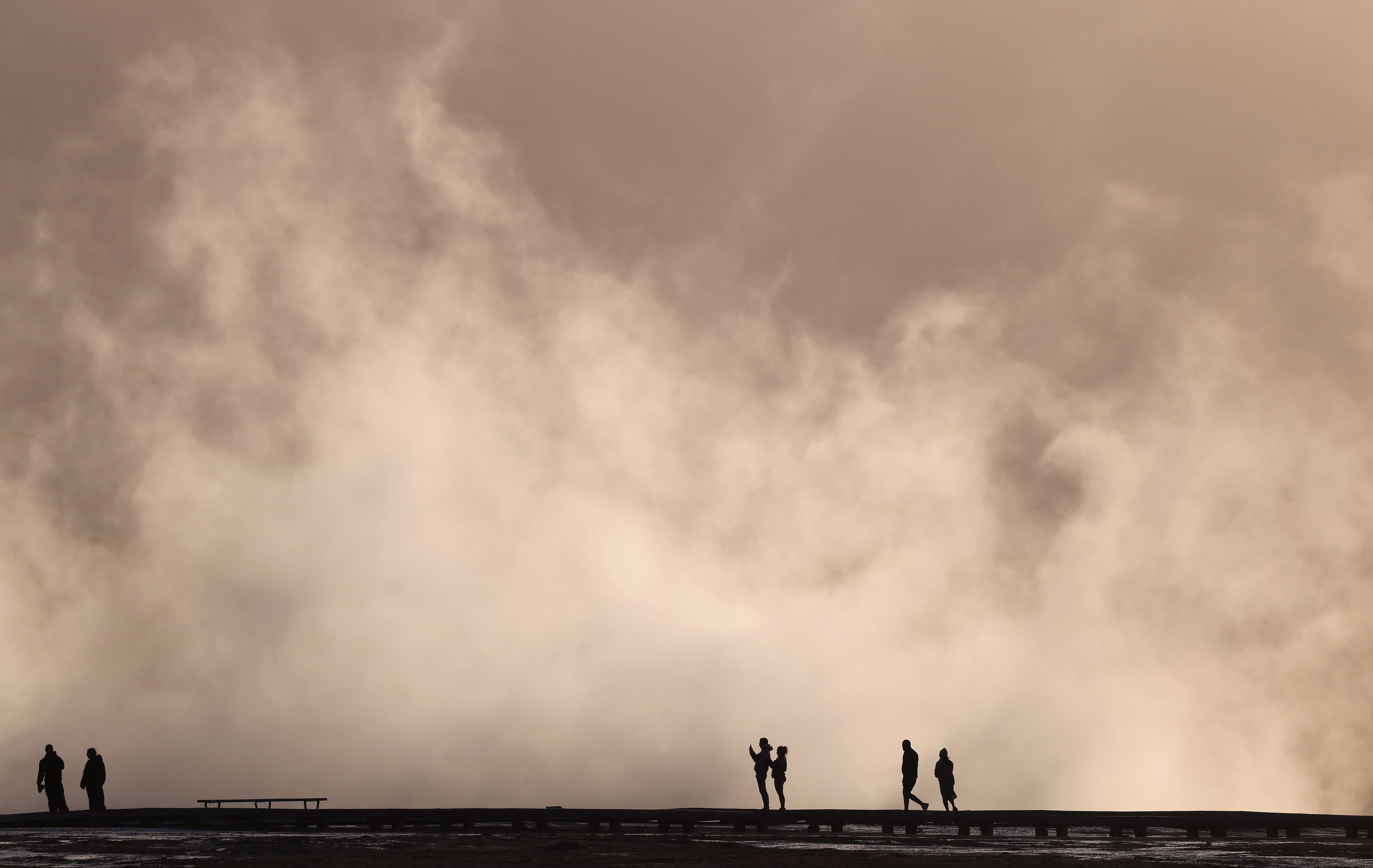 Several people are seen in silhouette, standing in front of large clouds of steam rising from a geyser basin.