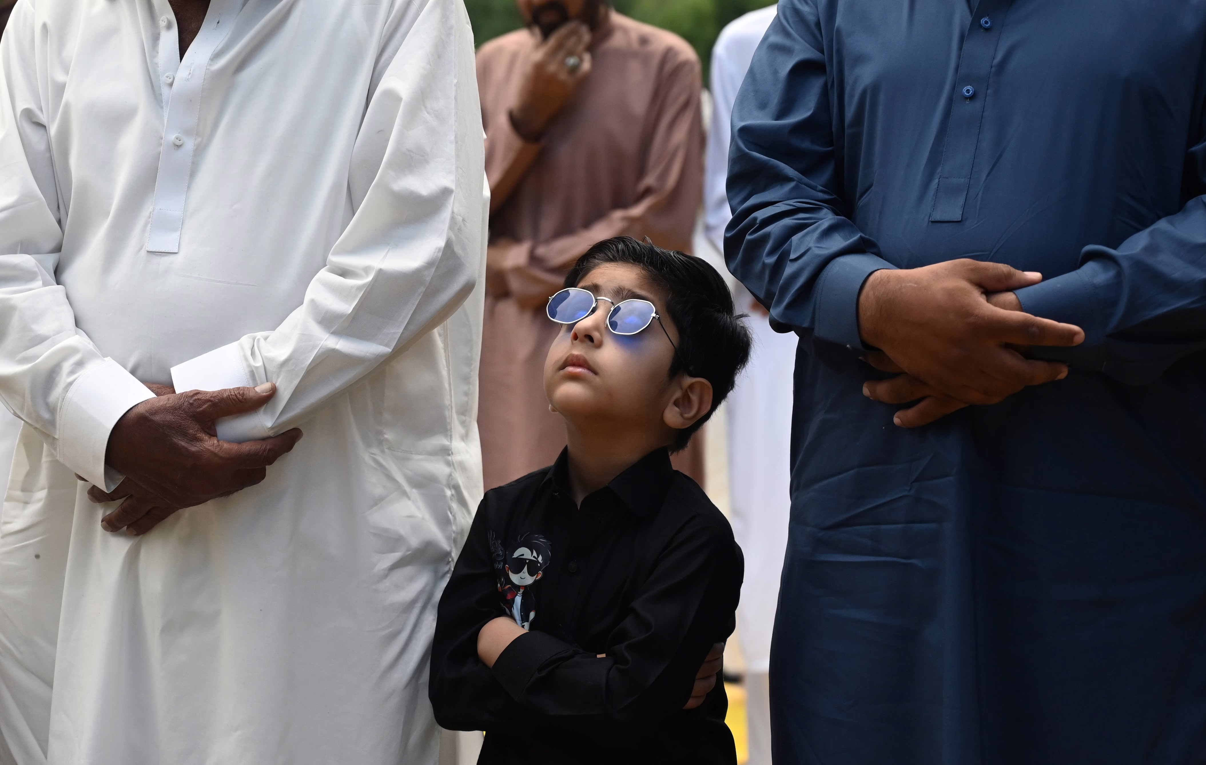 A boy wearing sunglasses looks up while standing beside adults during a Muslim prayer session.
