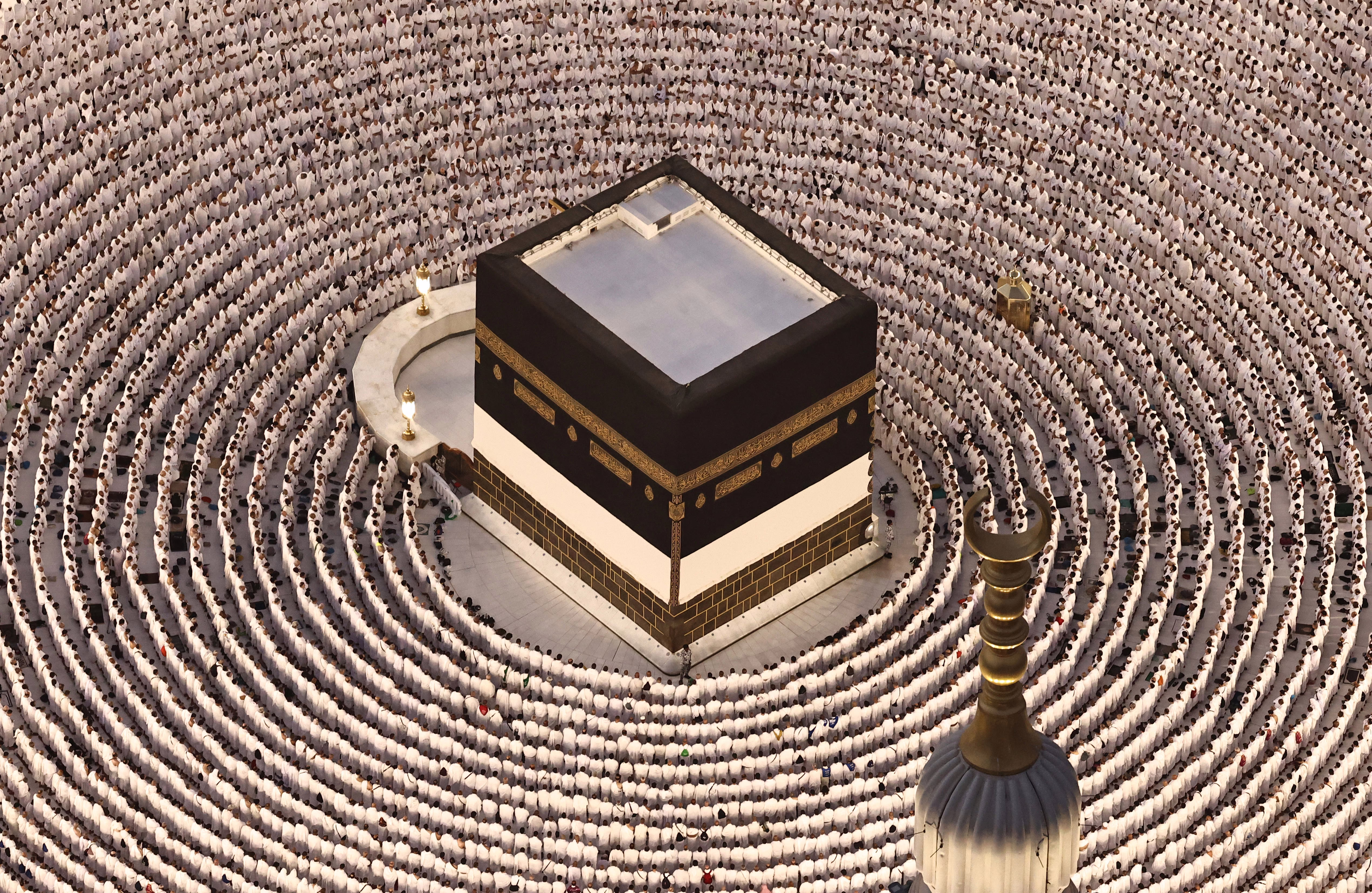 Many Muslim pilgrims dressed in white kneel in prayer, arranged in concentric circles around a tall cube-shaped shrine.