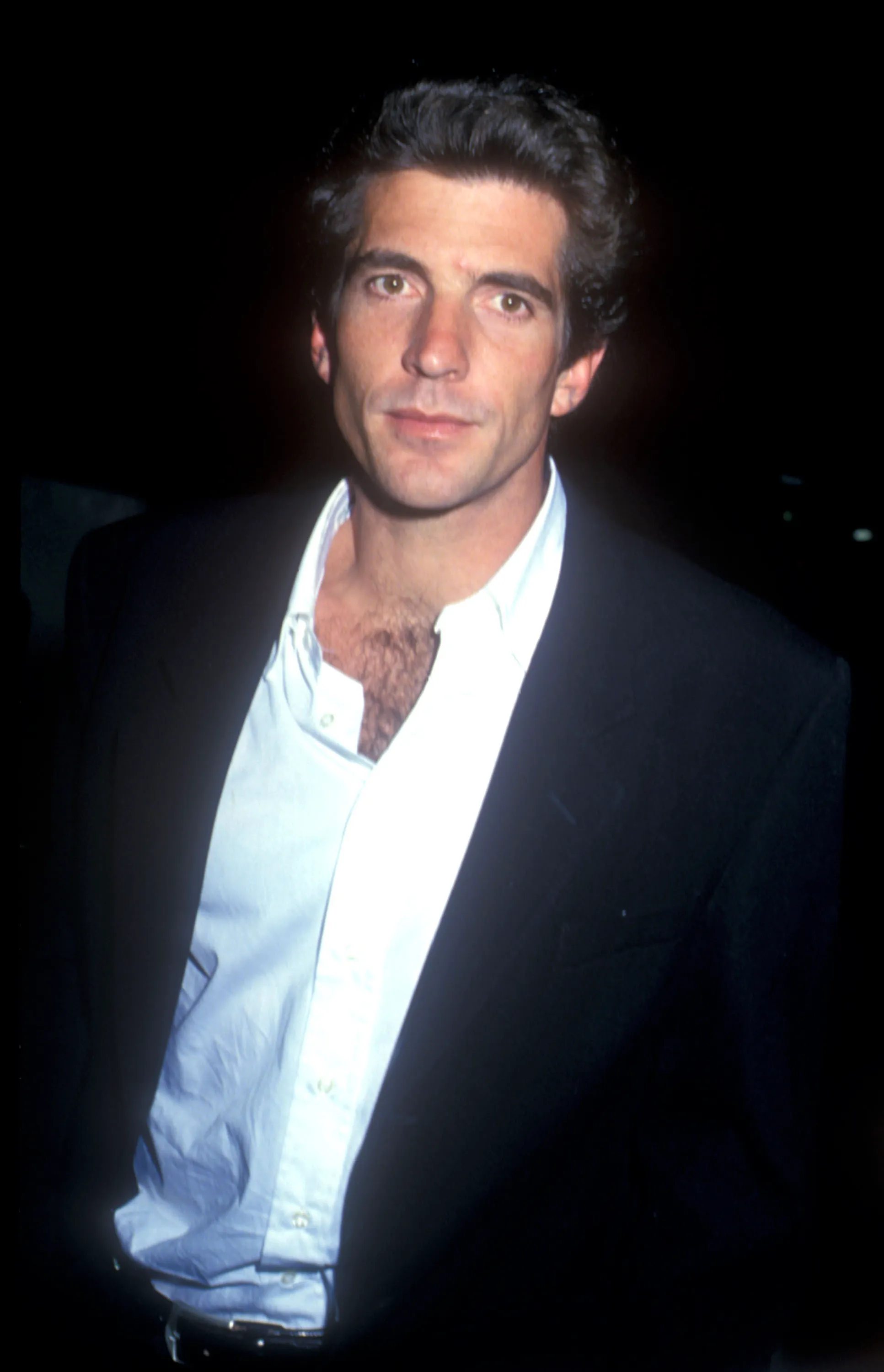 John F. Kennedy Jr. in 1993, looking forward with dark hair, a black jacket, and a partially unbuttoned white shirt.