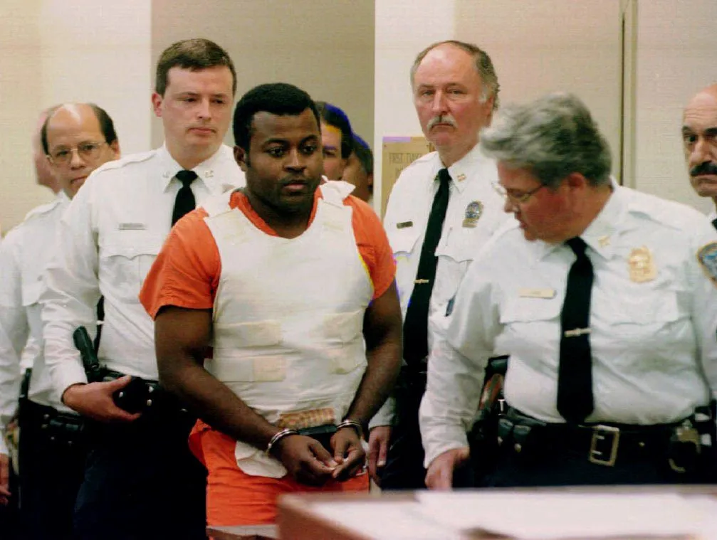 Colin Ferguson, wearing an orange jumpsuit and a white bulletproof vest, in handcuffs, surrounded by court officers during his initial hearing.